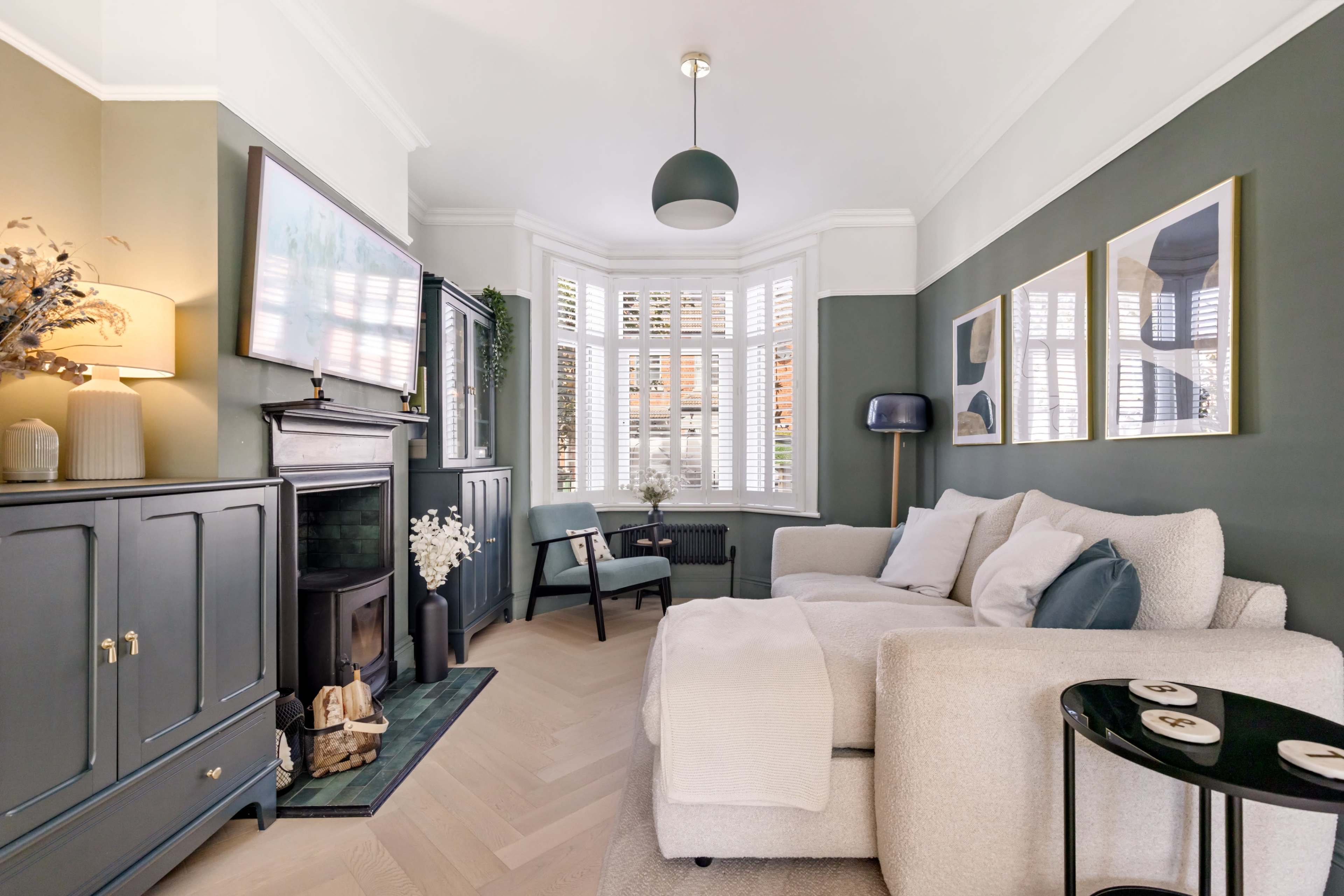 The image features a cozy living room with a light-colored sofa, a dark cabinet, and a bay window adorned with shutters.