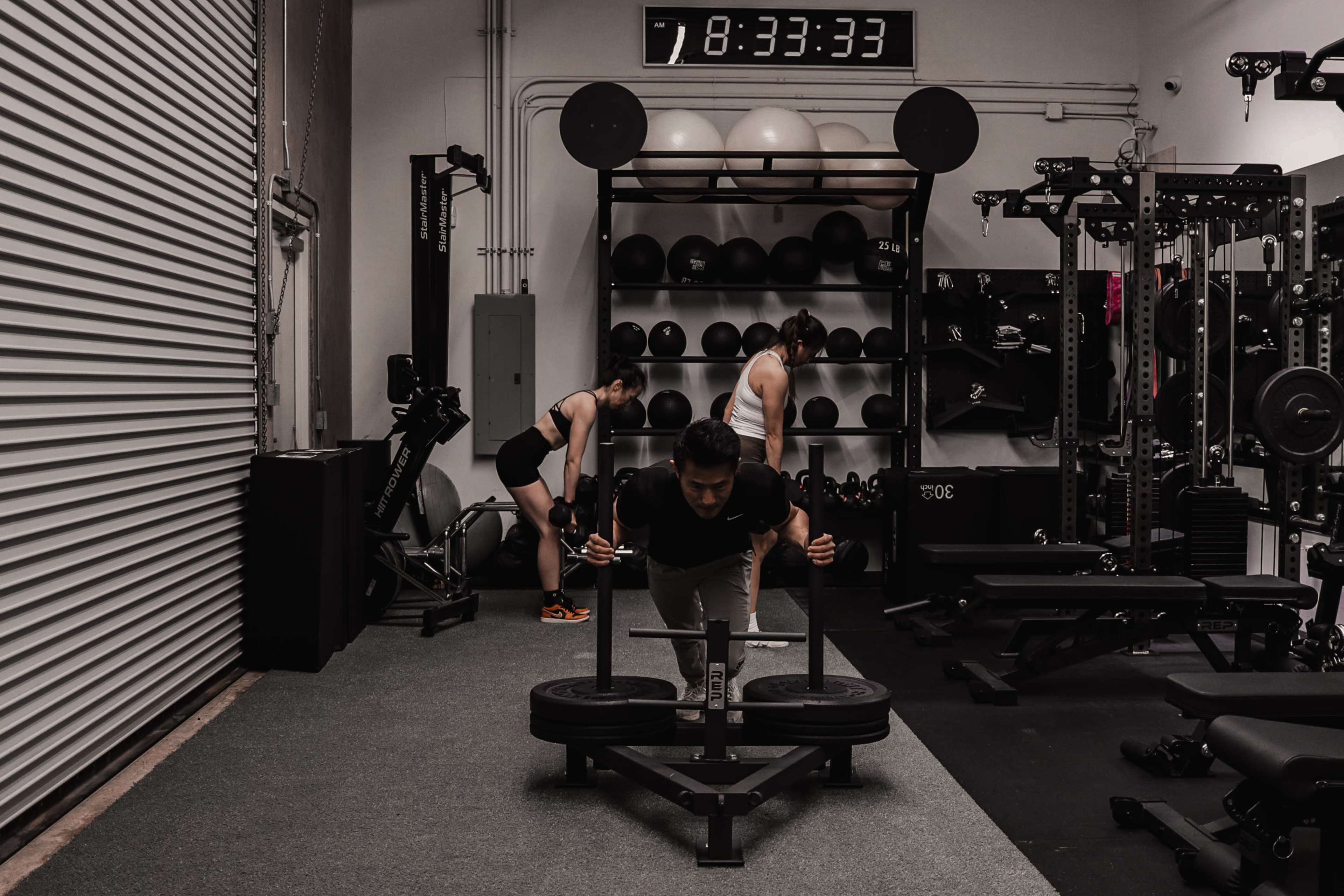 A person is pushing a weighted sled in a gym while two others are engaged in different exercises in the background.
