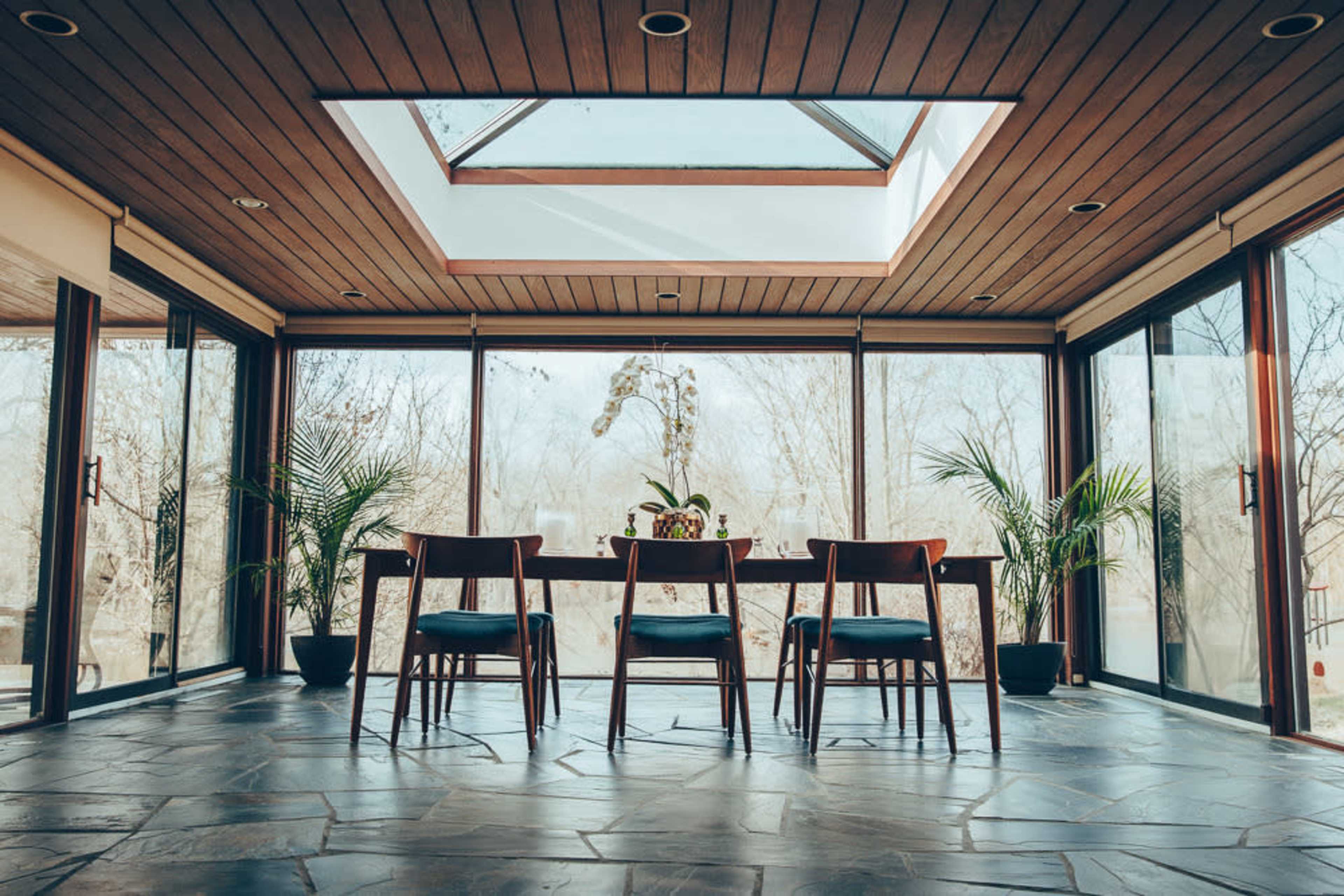 A modern dining area features a wooden table and chairs arranged under a skylight, surrounded by large glass windows and plants.