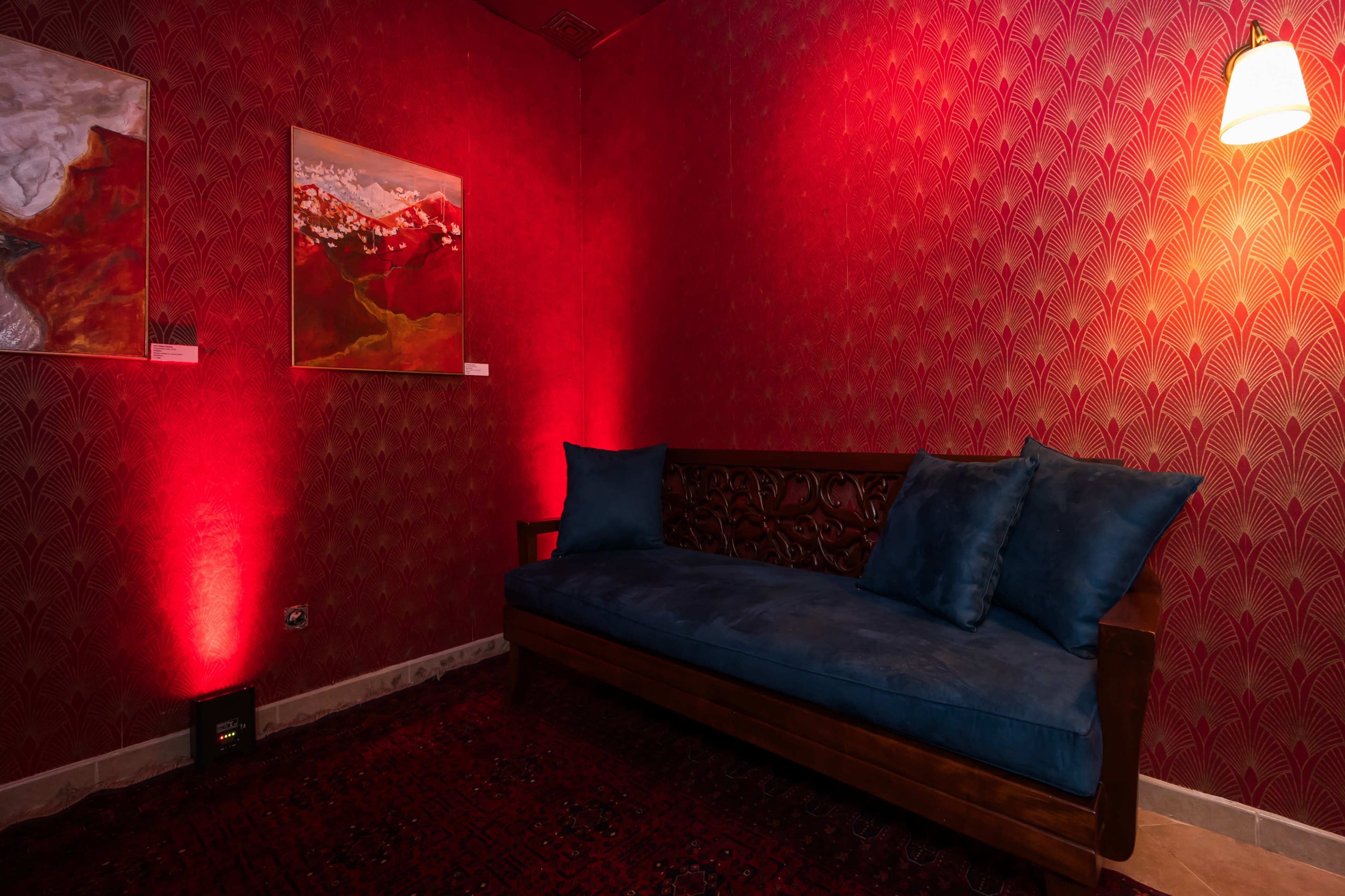 The image shows a small room with red patterned walls, featuring a wooden couch adorned with blue pillows and illuminated by soft lighting.
