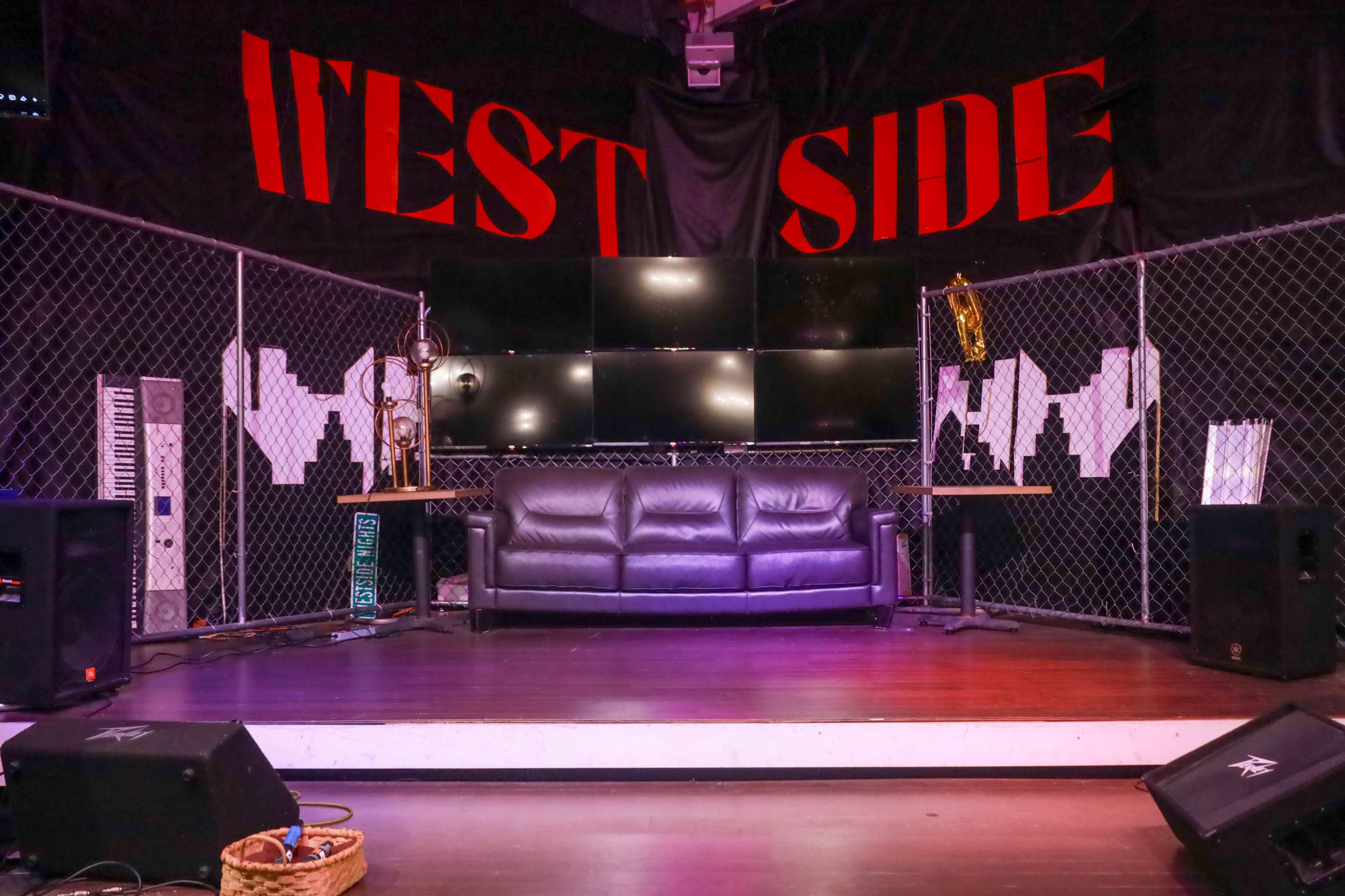 The image shows a stage area with a black couch, musical instruments, and multiple screens mounted on a backdrop featuring the phrase "WEST SIDE" in bold red letters.