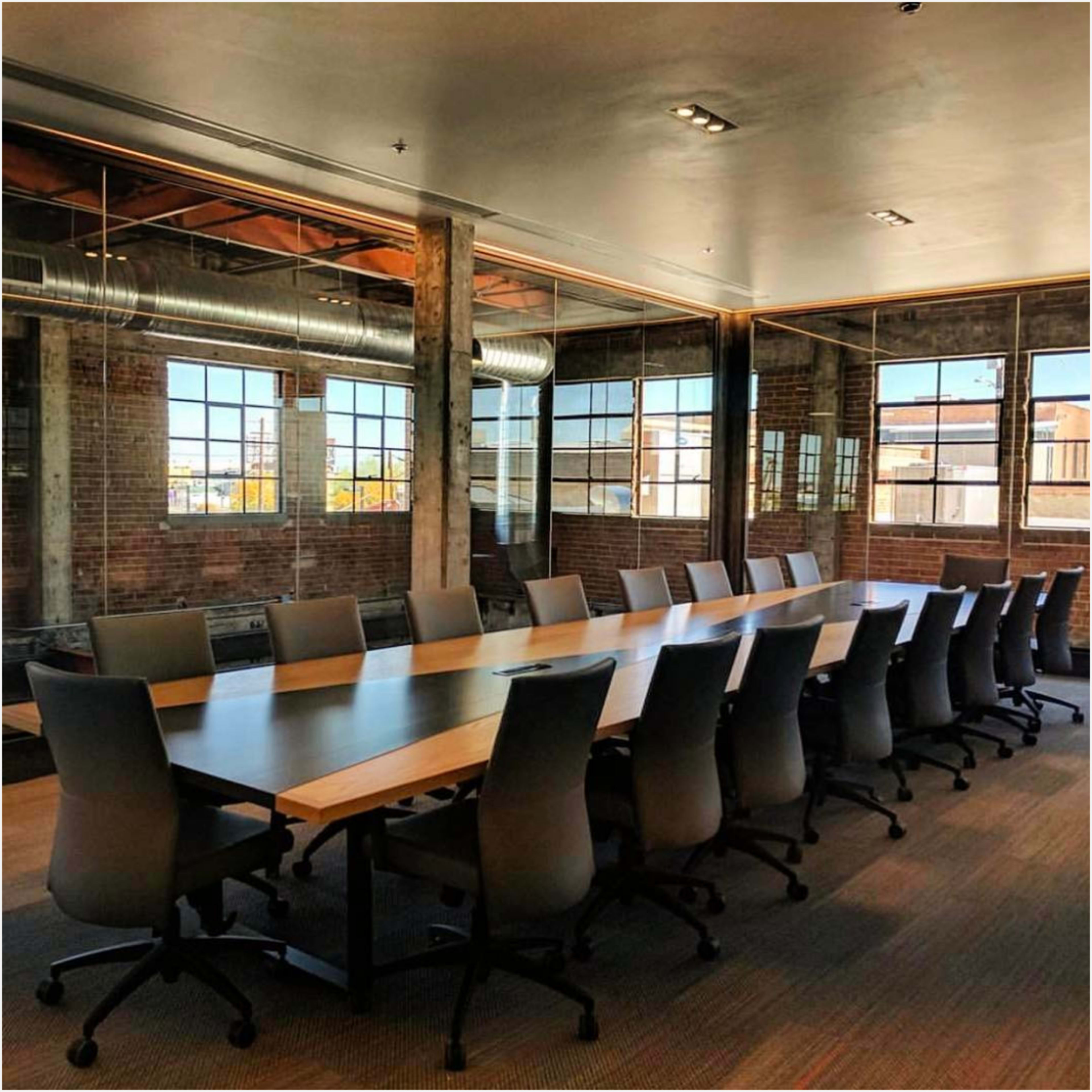A long conference table with ergonomic chairs is set up in a modern meeting room with large windows and exposed brick walls.