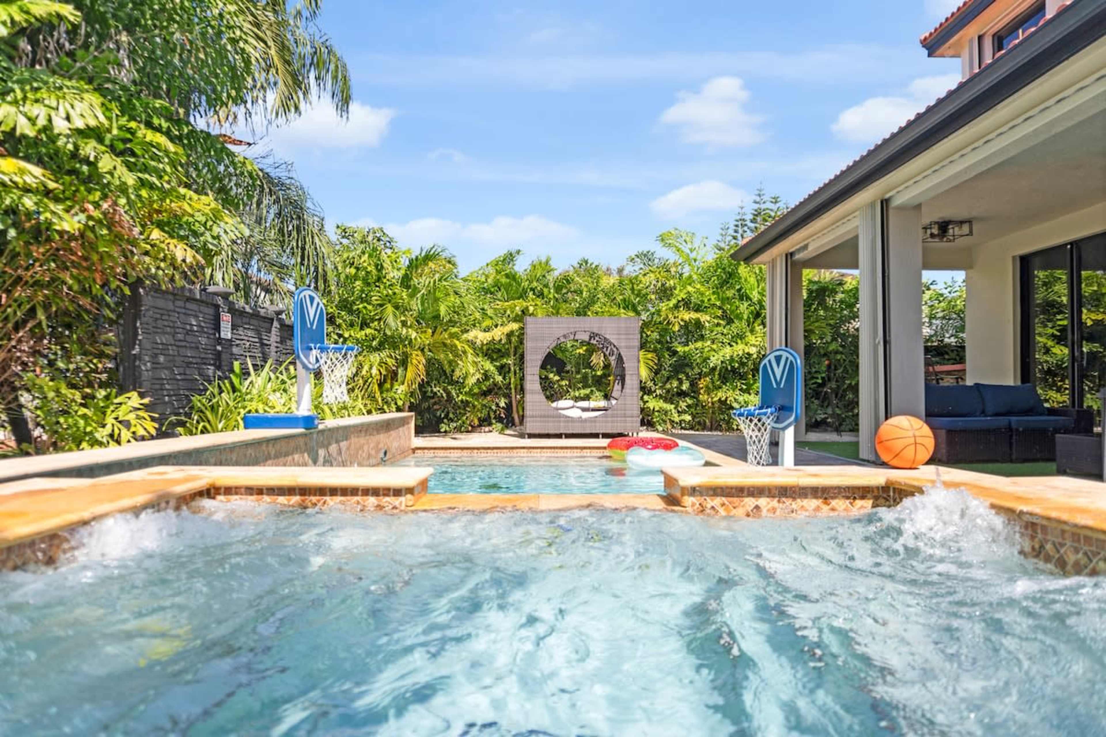 The image shows a backyard with a swimming pool featuring basketball hoops on the edges, surrounded by lush tropical greenery and a modern seating area in the background.