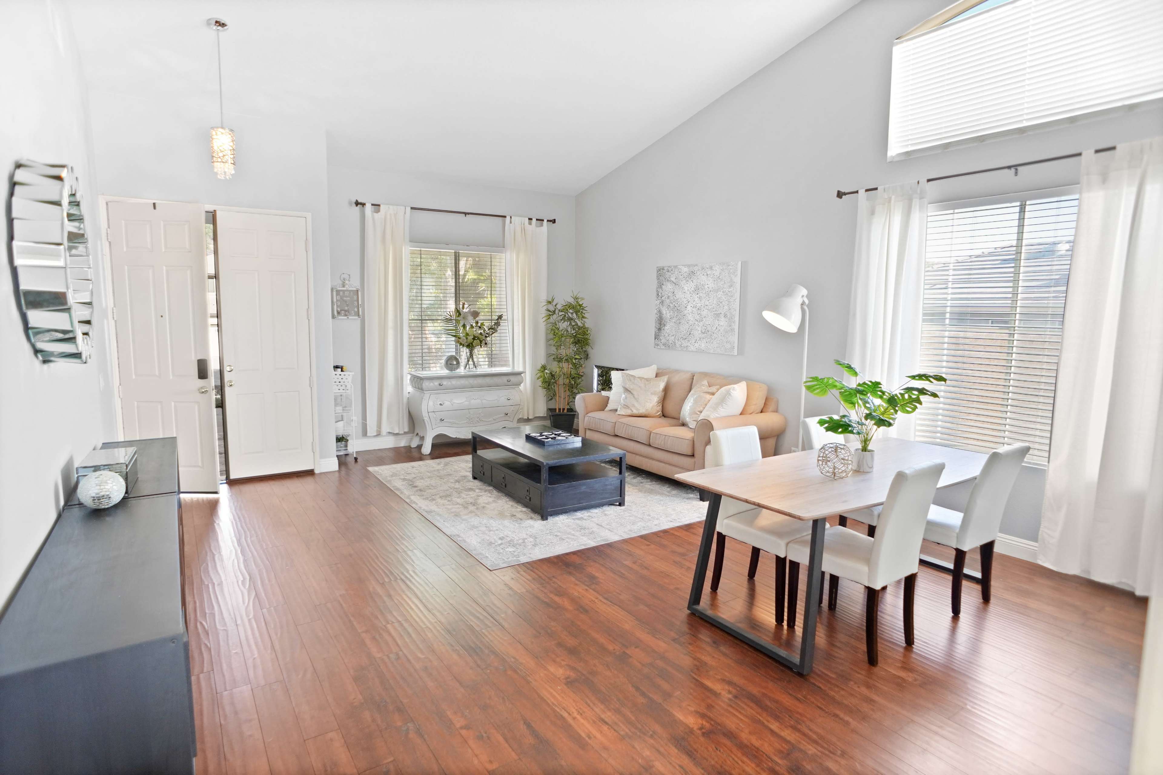 The image shows a modern living room with a sofa, a coffee table, a dining area, and large windows allowing natural light to enter.