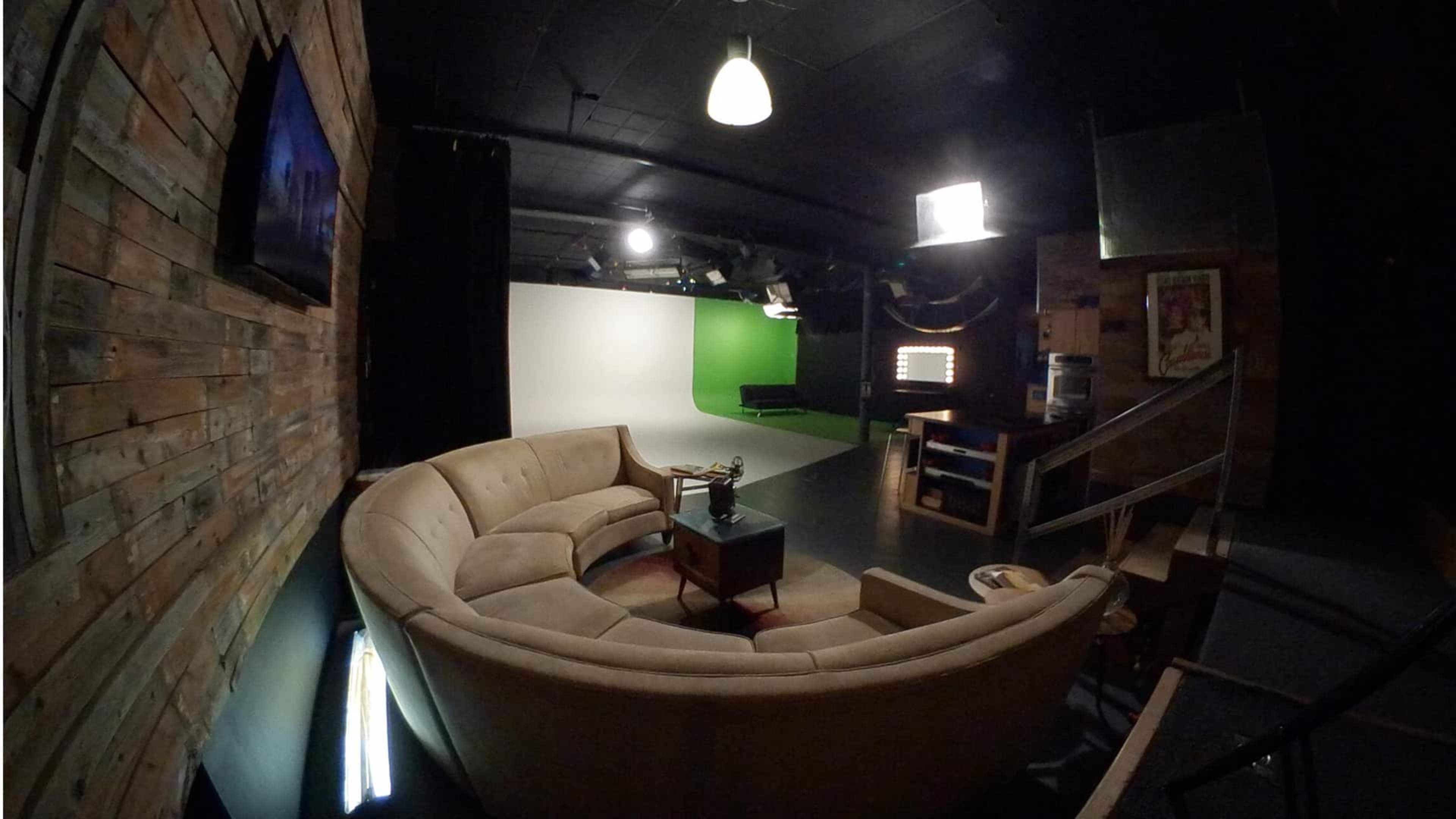 A studio space featuring a curved beige couch, a coffee table, and a backdrop with green screen and light fixtures.