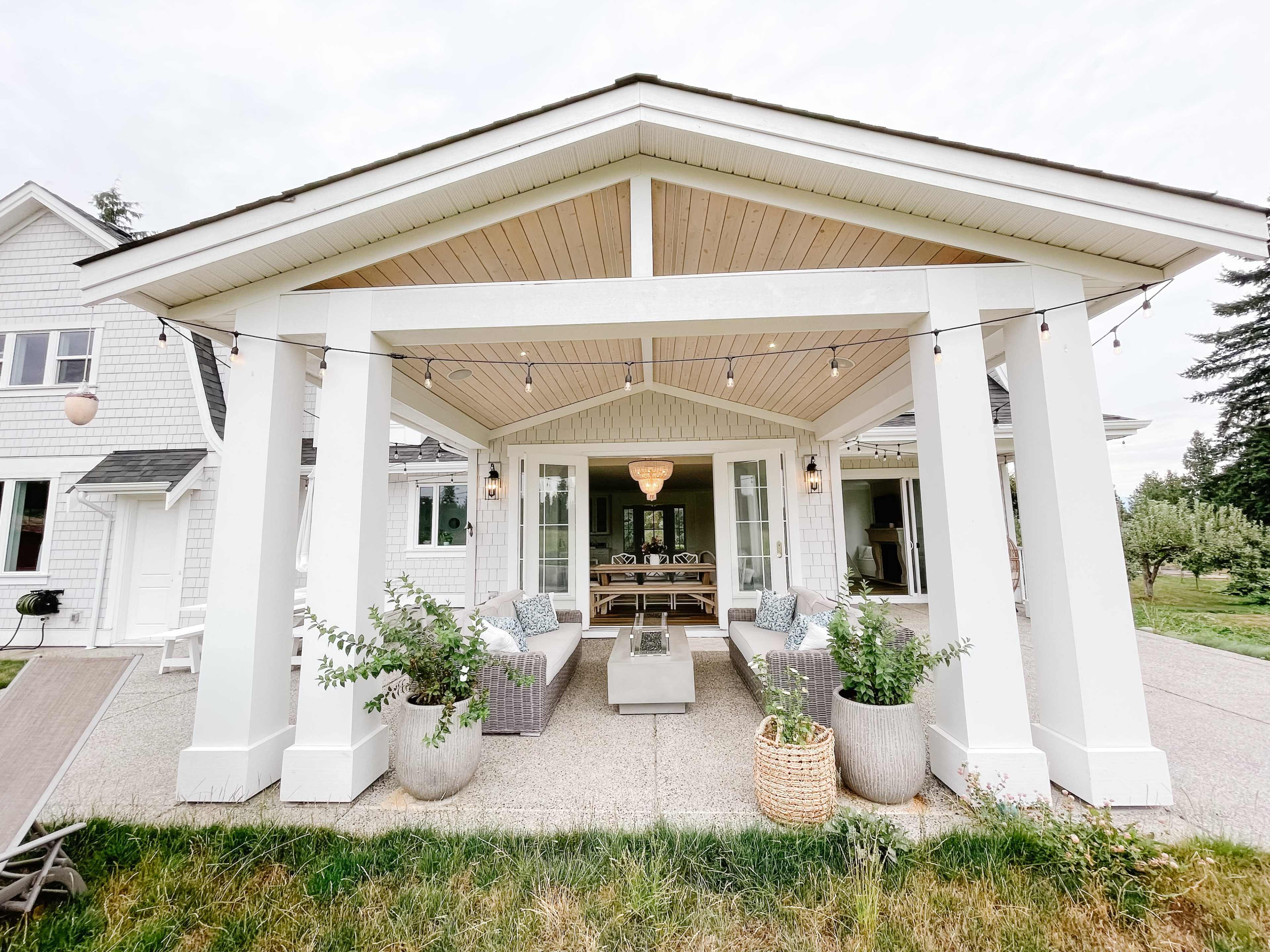A covered outdoor patio with large white columns, seating arrangements, and potted plants, leading to a house in the background.