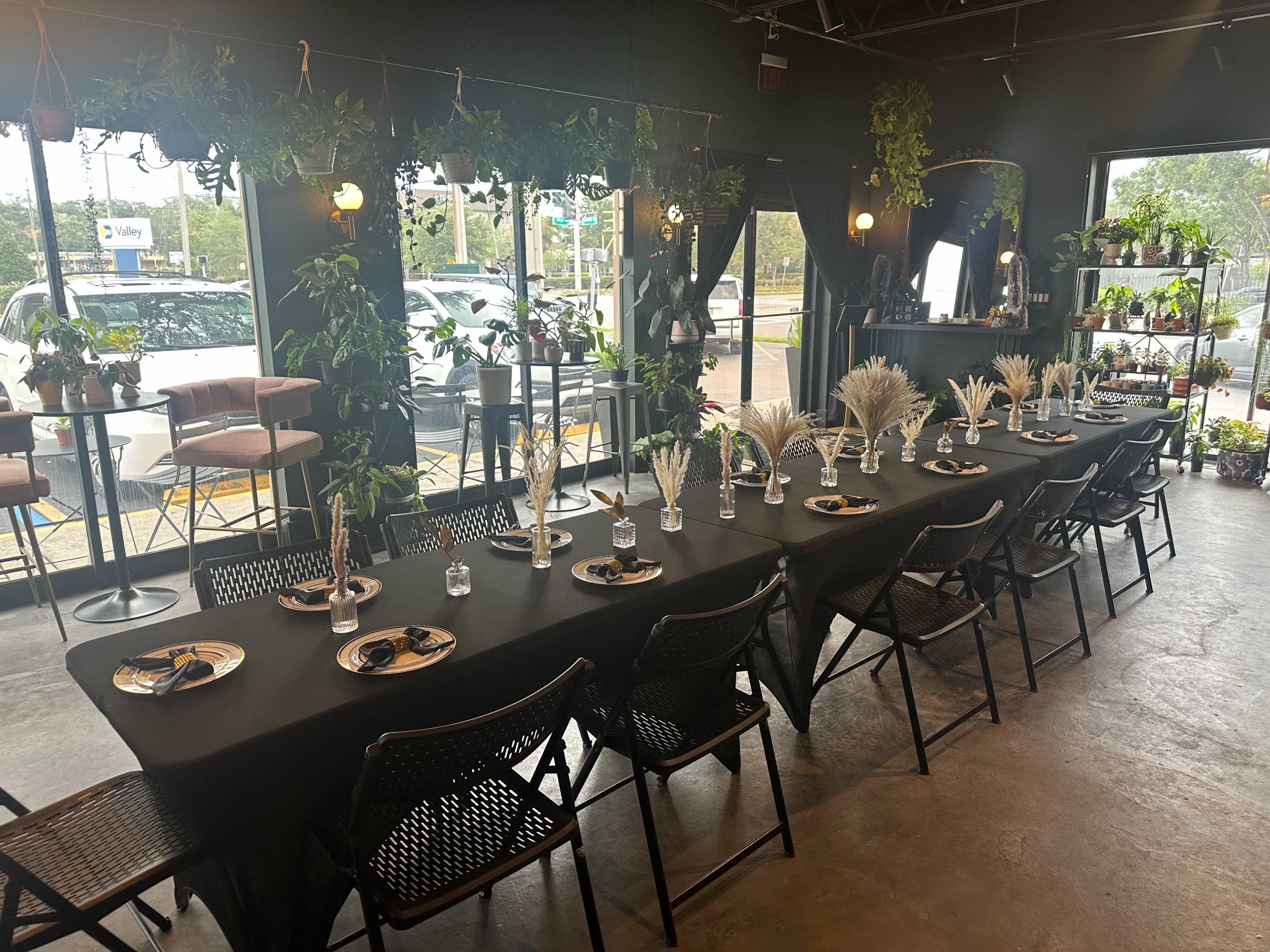 A long dining table is set with black tablecloths and decorative arrangements, surrounded by plants and large windows, creating a vibrant indoor space.