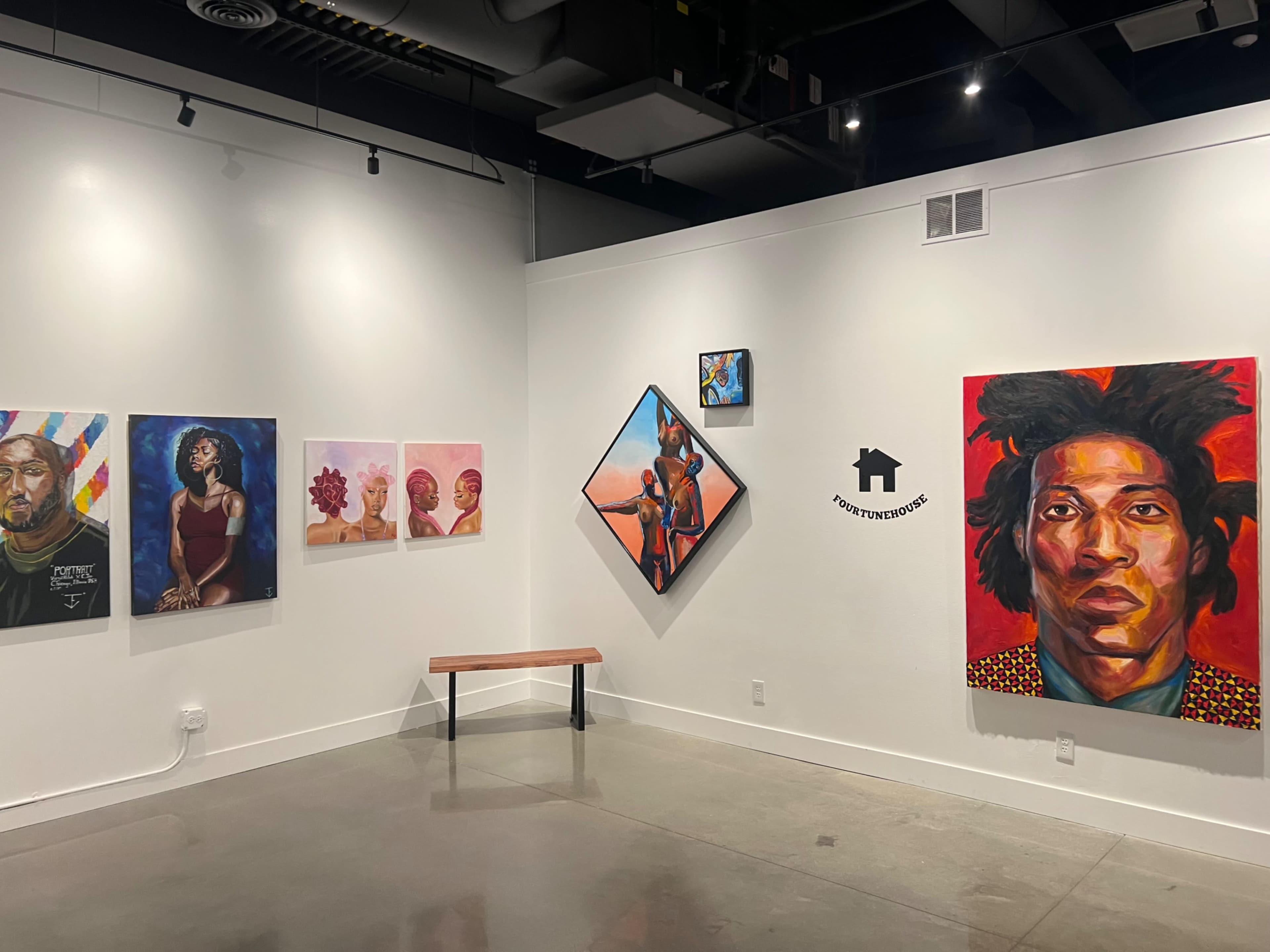 An art gallery wall displaying a variety of artwork, including portraits and abstract pieces, with a bench in front for visitors.