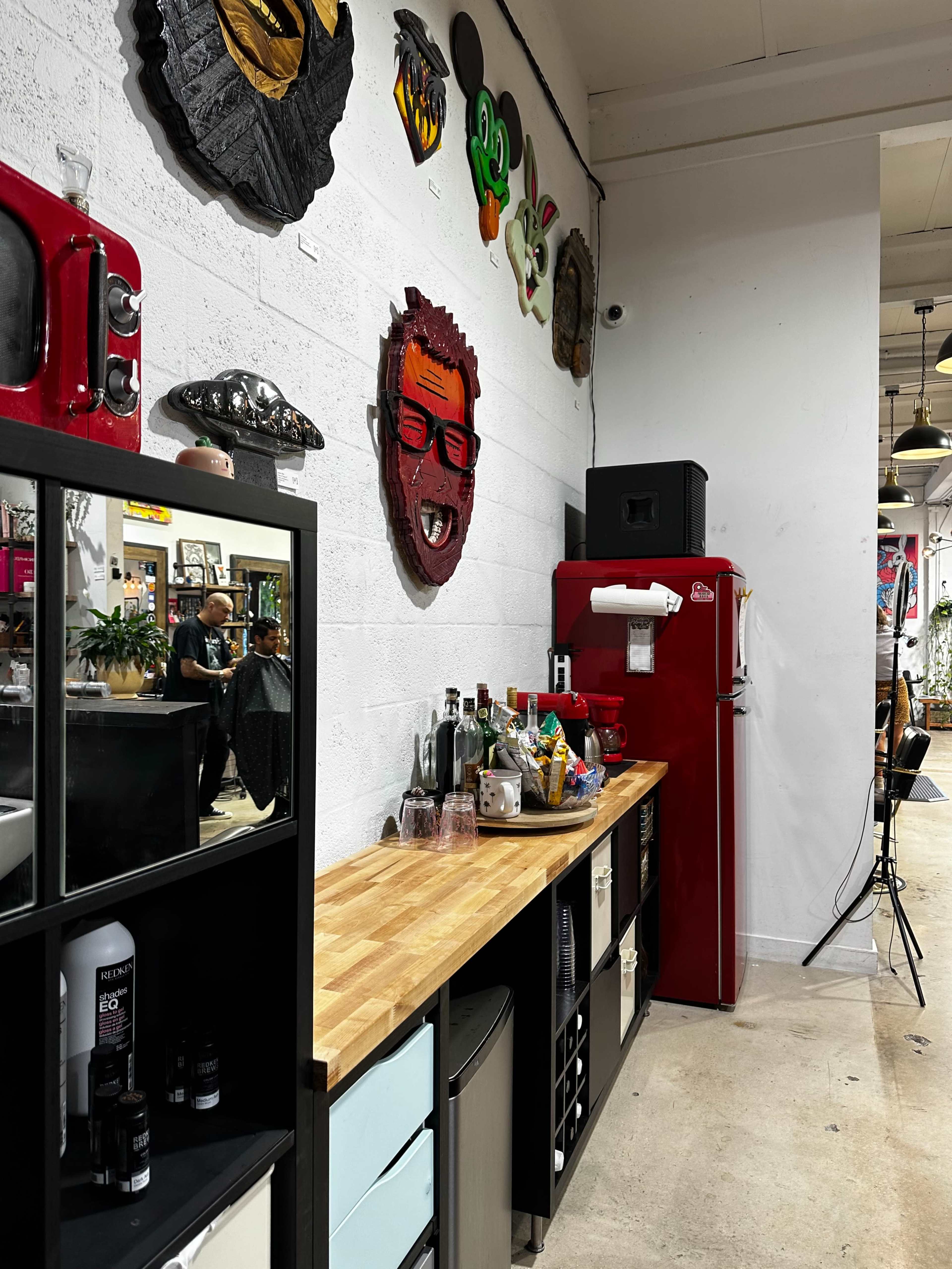 The image shows a vibrant interior space featuring a wooden countertop, a red refrigerator, various decorative masks on the wall, and a shelf with bottles and glasses.