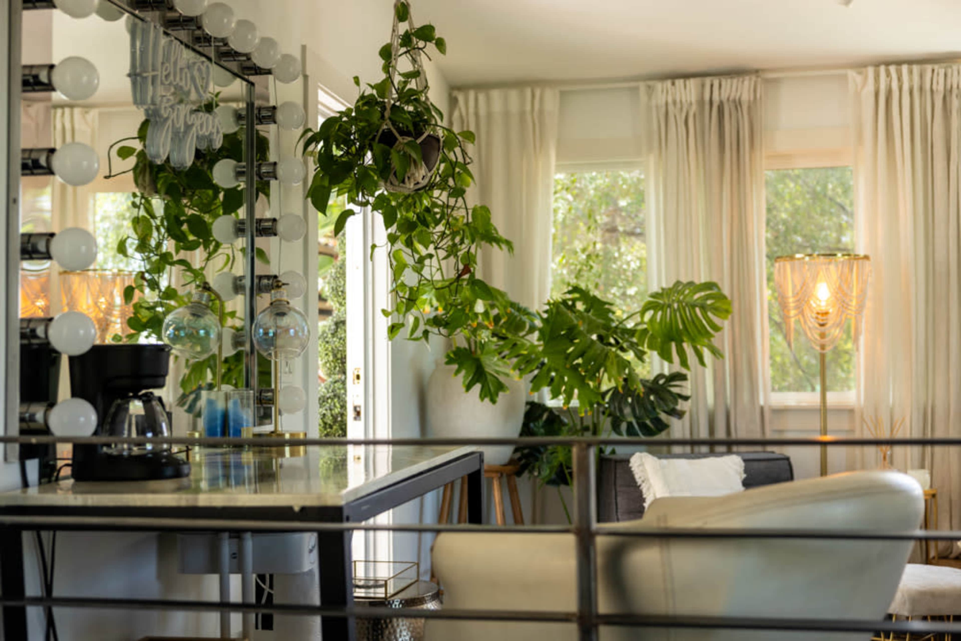 The image shows a bright living space with a plant-filled corner, a coffee station, and a large mirror with light bulbs.