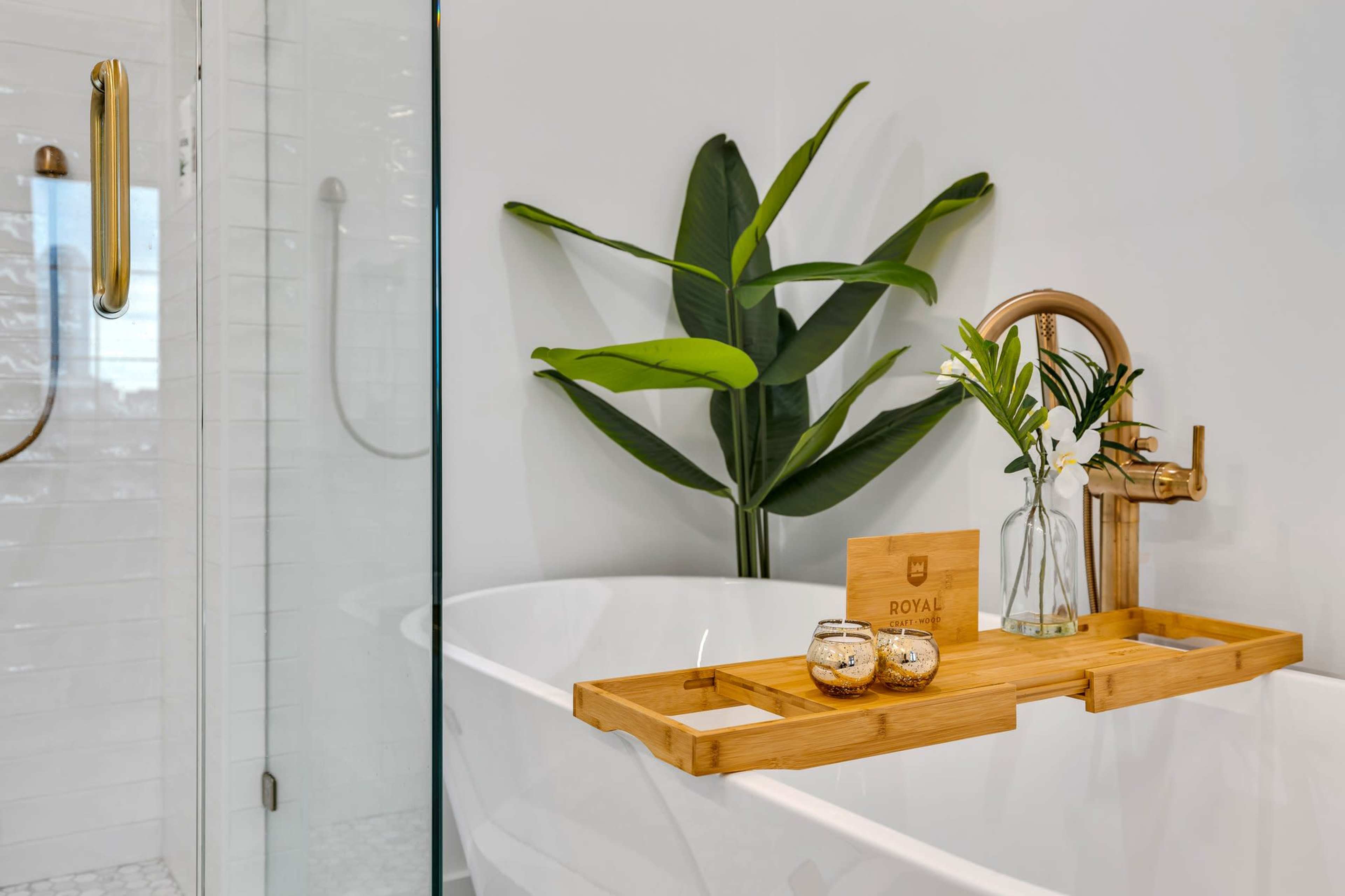 A bamboo bath tray holds decorative items and a small vase with flowers beside a freestanding bathtub and a glass shower enclosure.