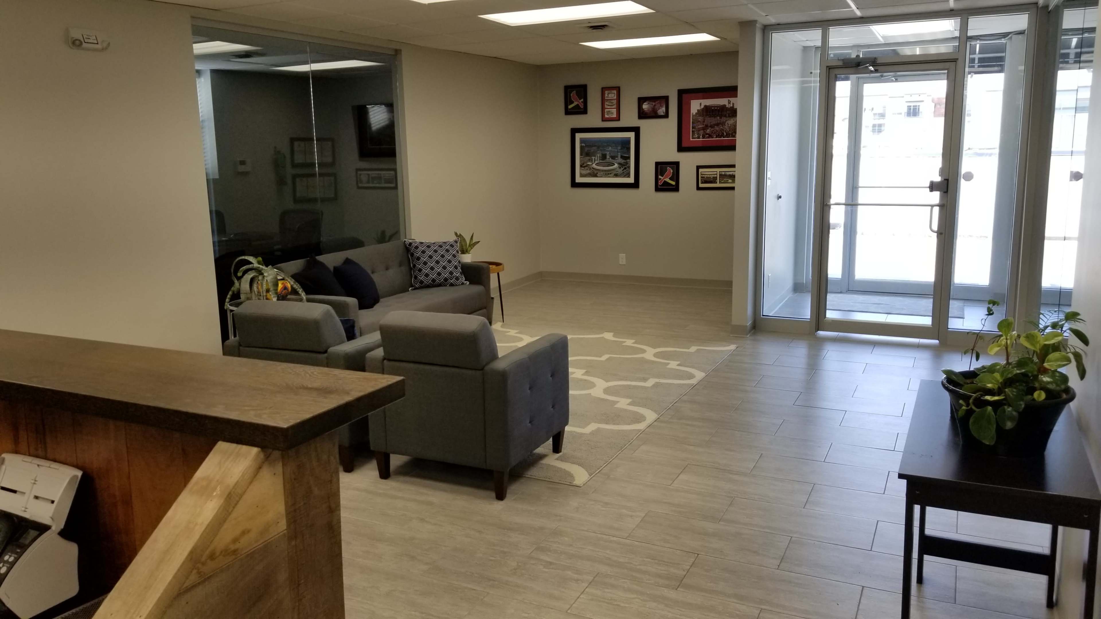 The image shows a bright lobby area with gray seating, a geometric rug, and glass doors leading outside.
