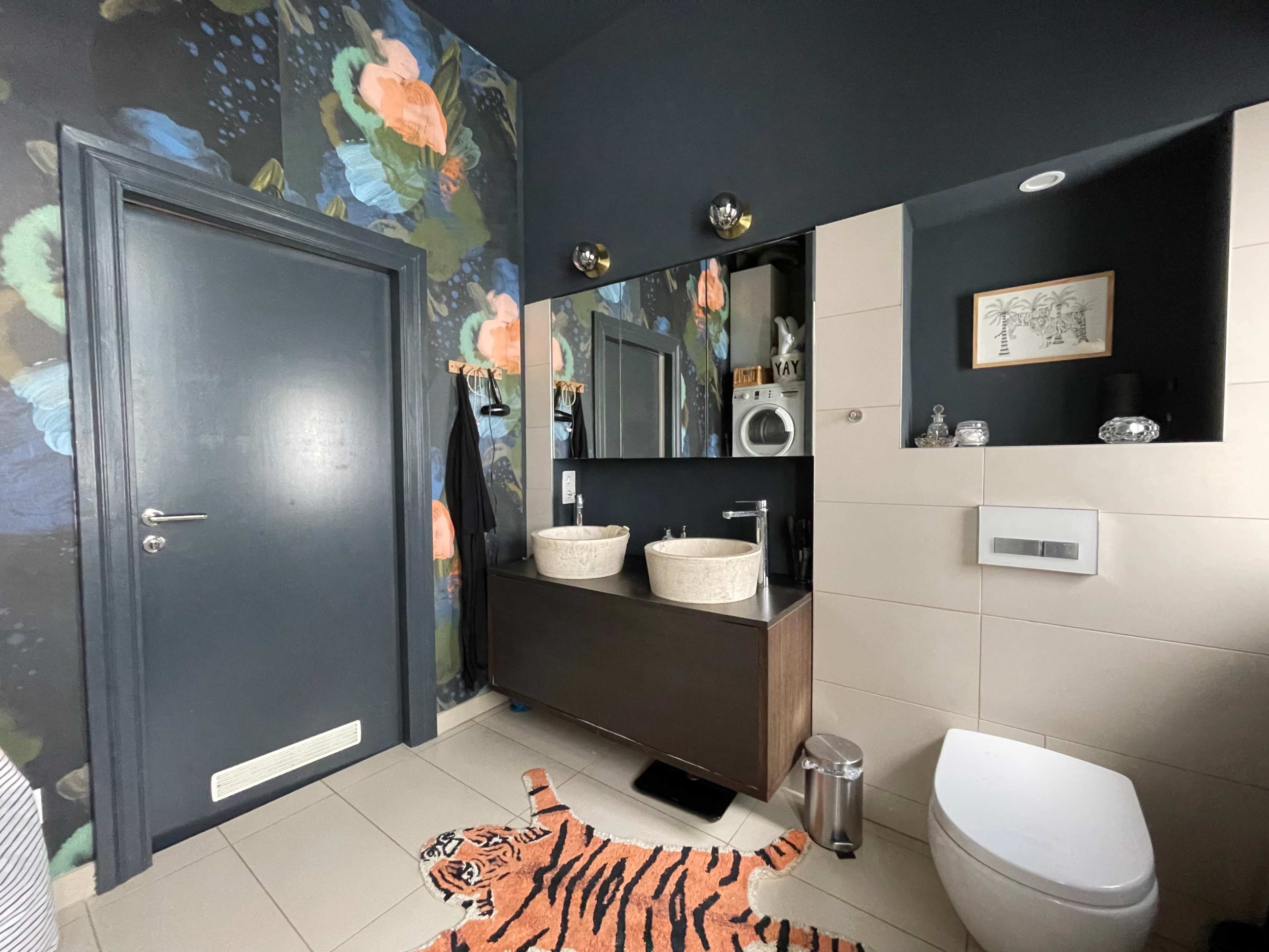 The bathroom features a dark floral wallpaper, a wooden vanity with two round sinks, and a tiger-patterned rug on the floor.