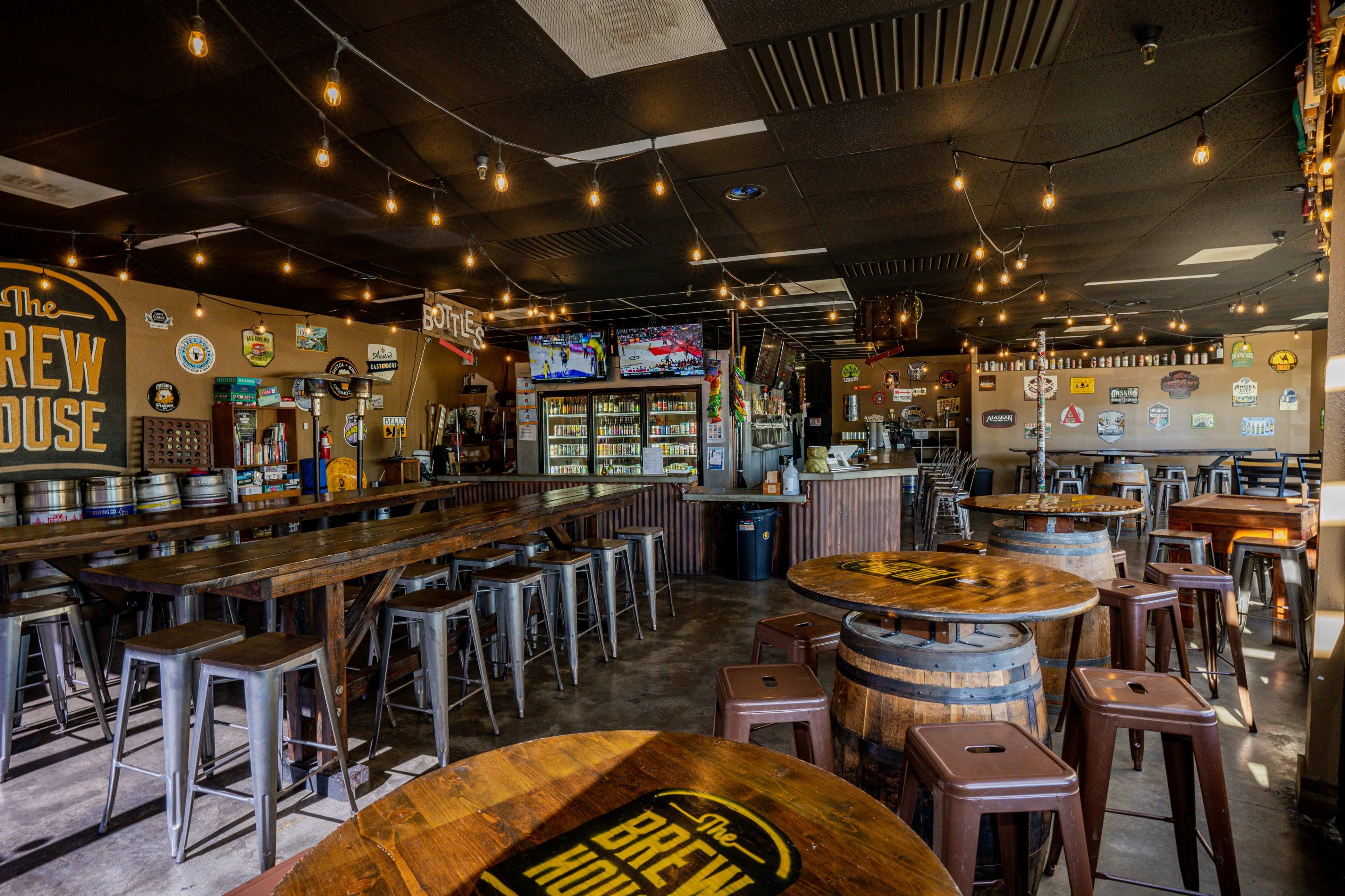 The interior of a brew house features wooden tables, metal stools, a bar with various beverages, and string lights hanging from the ceiling.