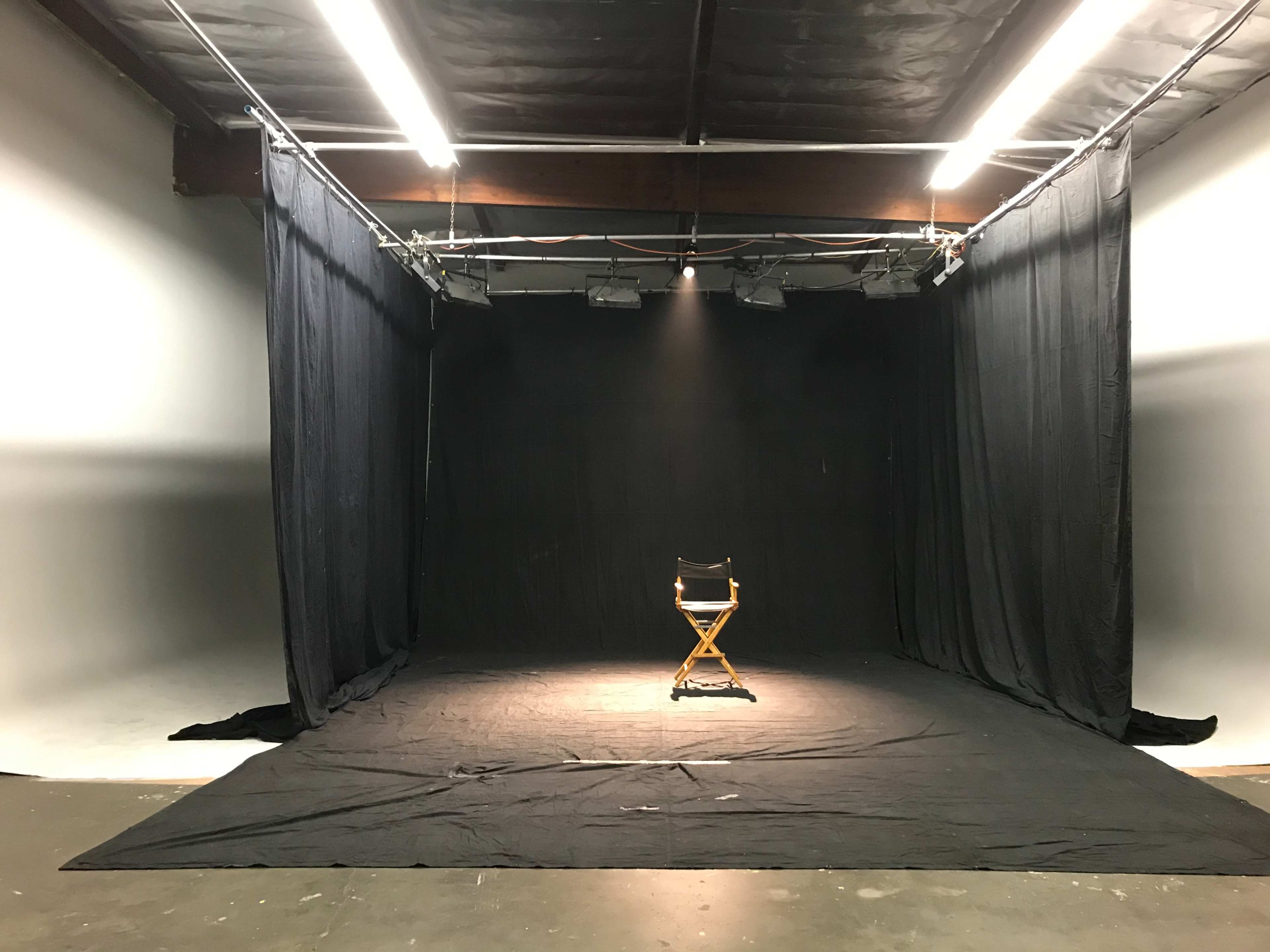 A spotlight illuminates an empty director's chair in a dimly lit studio surrounded by black curtains.