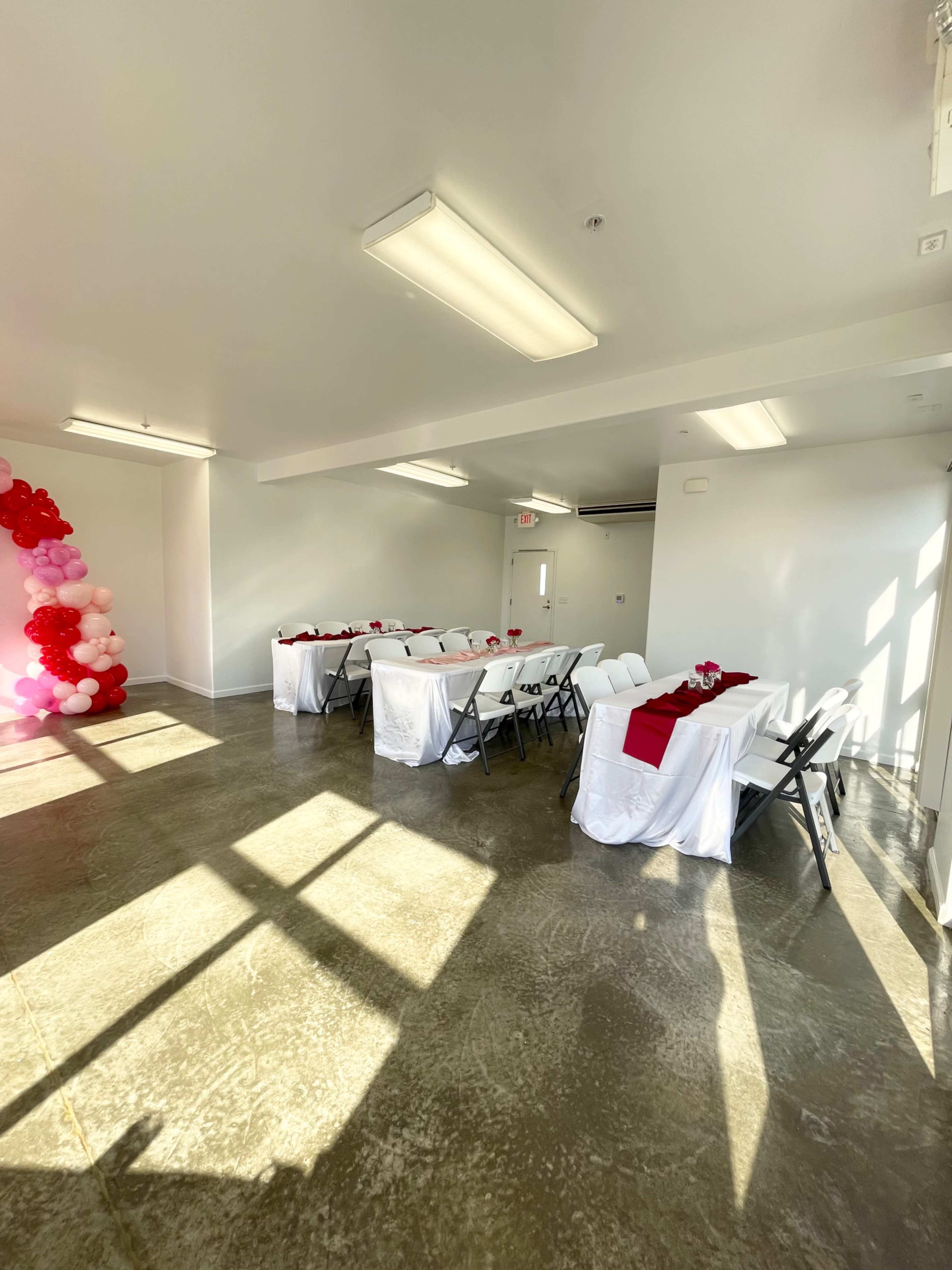 A room with two tables set for an event, each covered with white cloths and adorned with red accents, illuminated by natural light streaming through windows.