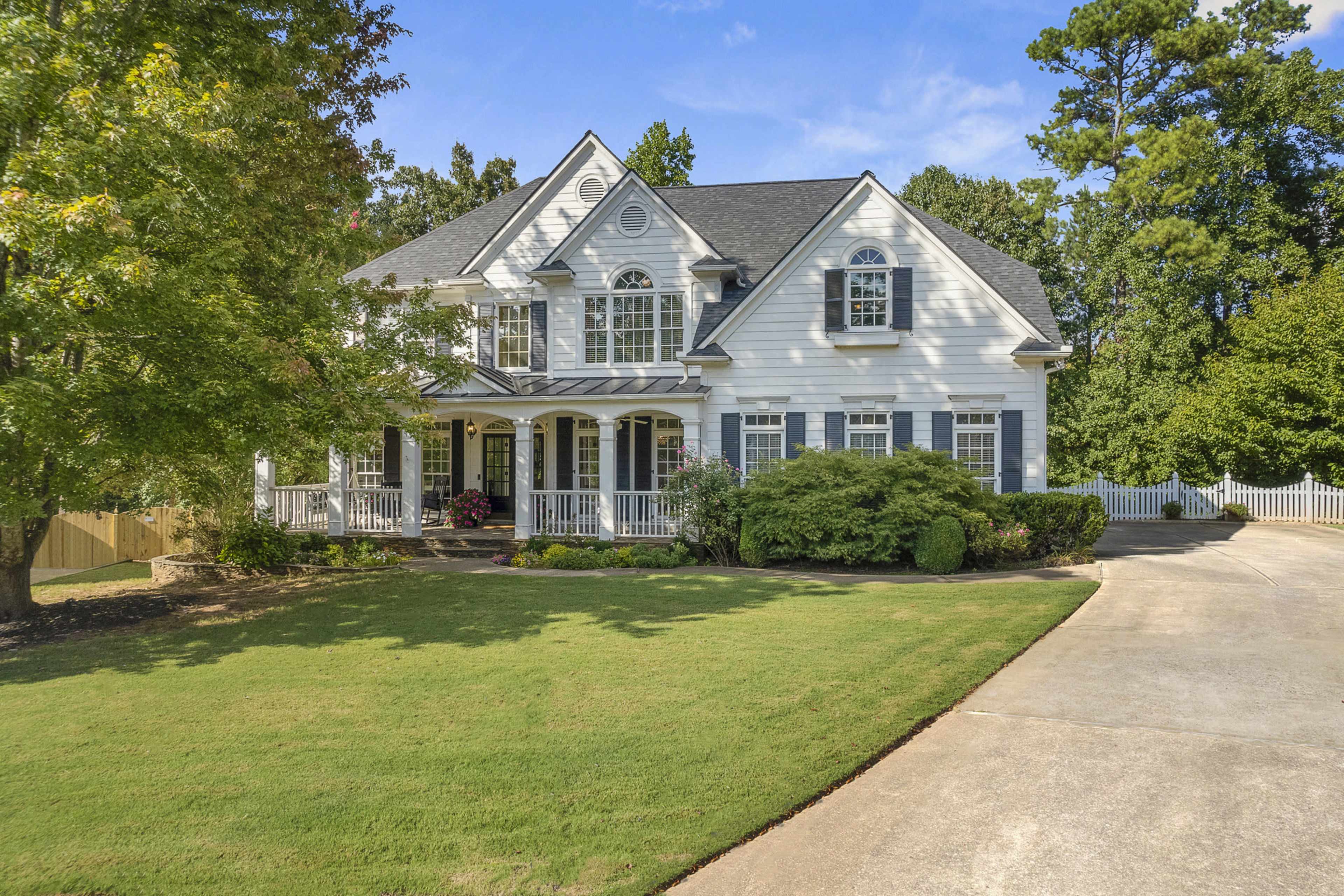A two-story white house with a covered porch and dark shutters is set amidst trees on a grassy lawn, with a paved driveway leading up to it.