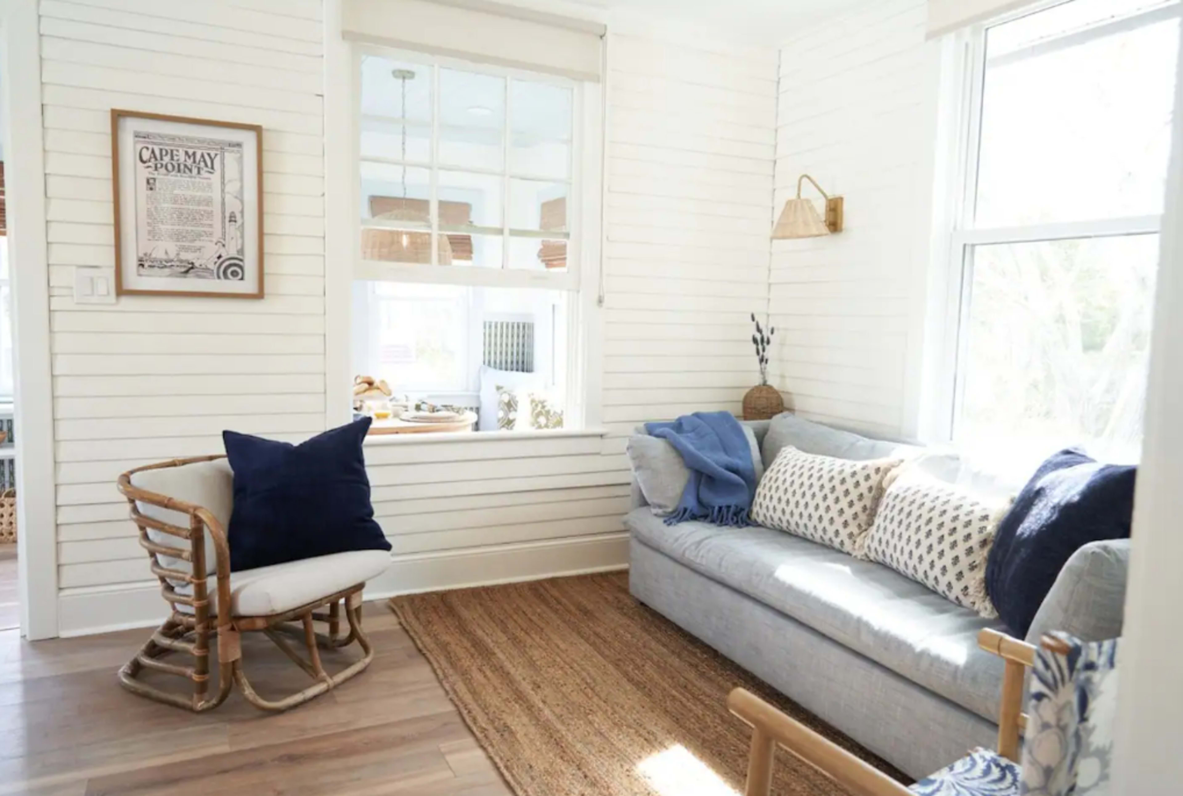 A light-colored living room features a blue sofa, a rocking chair, and large windows with natural light illuminating the space.