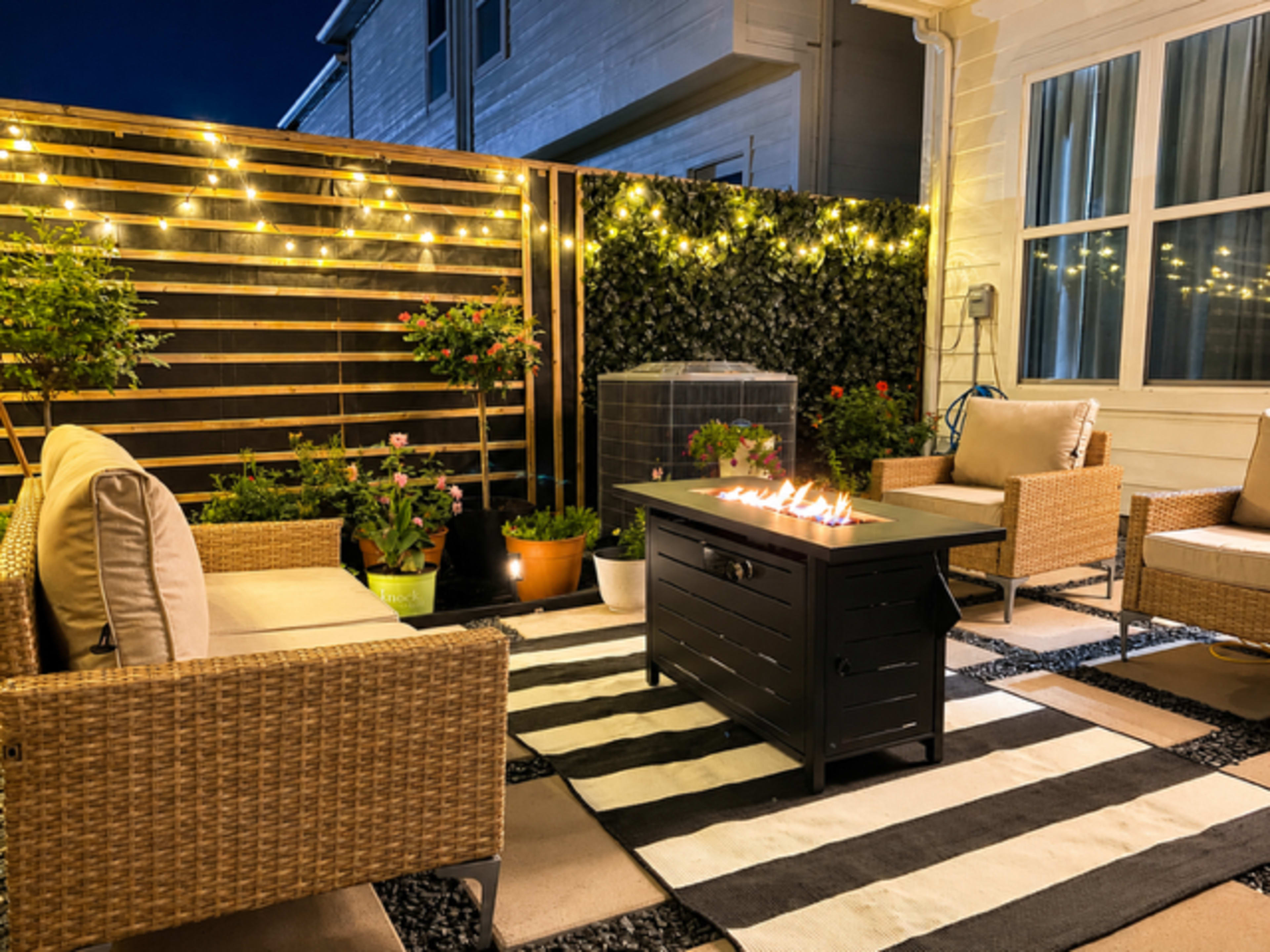 The image shows a cozy outdoor seating area with two wicker chairs, a black fire table, potted plants, and string lights illuminating a patio at night.