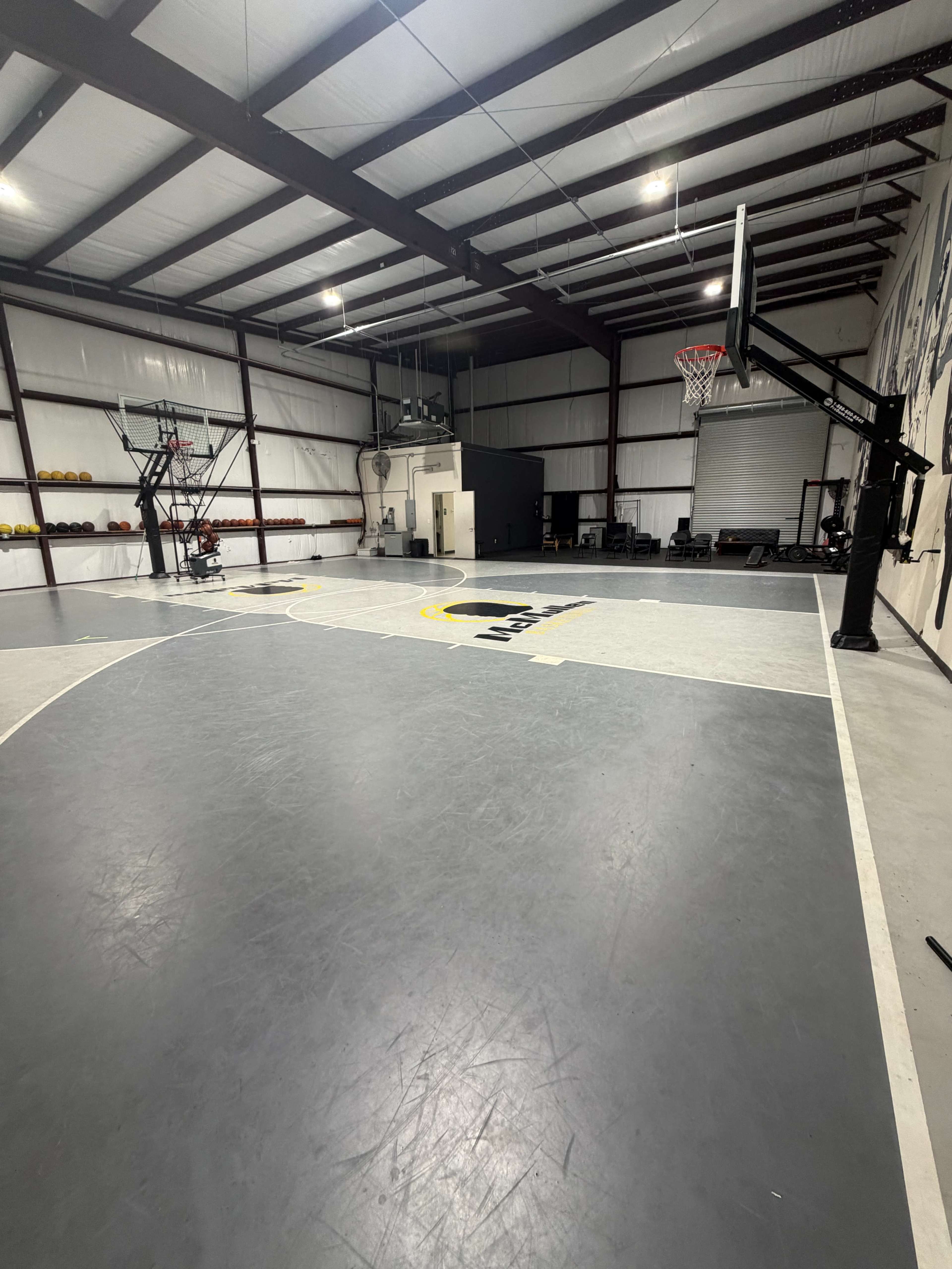 The image shows an indoor basketball court with a gray floor, a basketball hoop, and training equipment in a spacious facility.