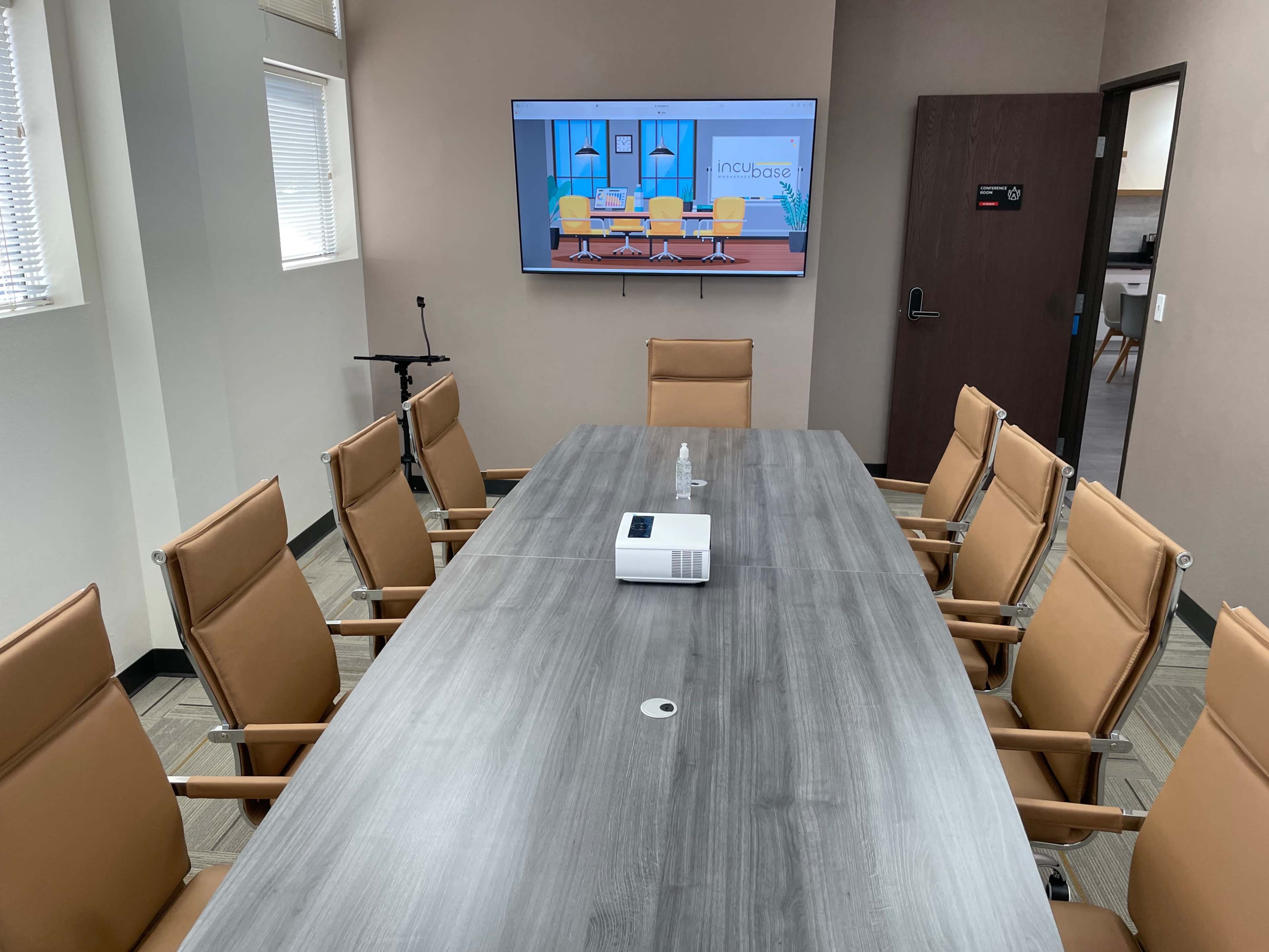 A conference room features a long wooden table surrounded by eight leather chairs, with a projector and a screen displaying a graphic on the wall.