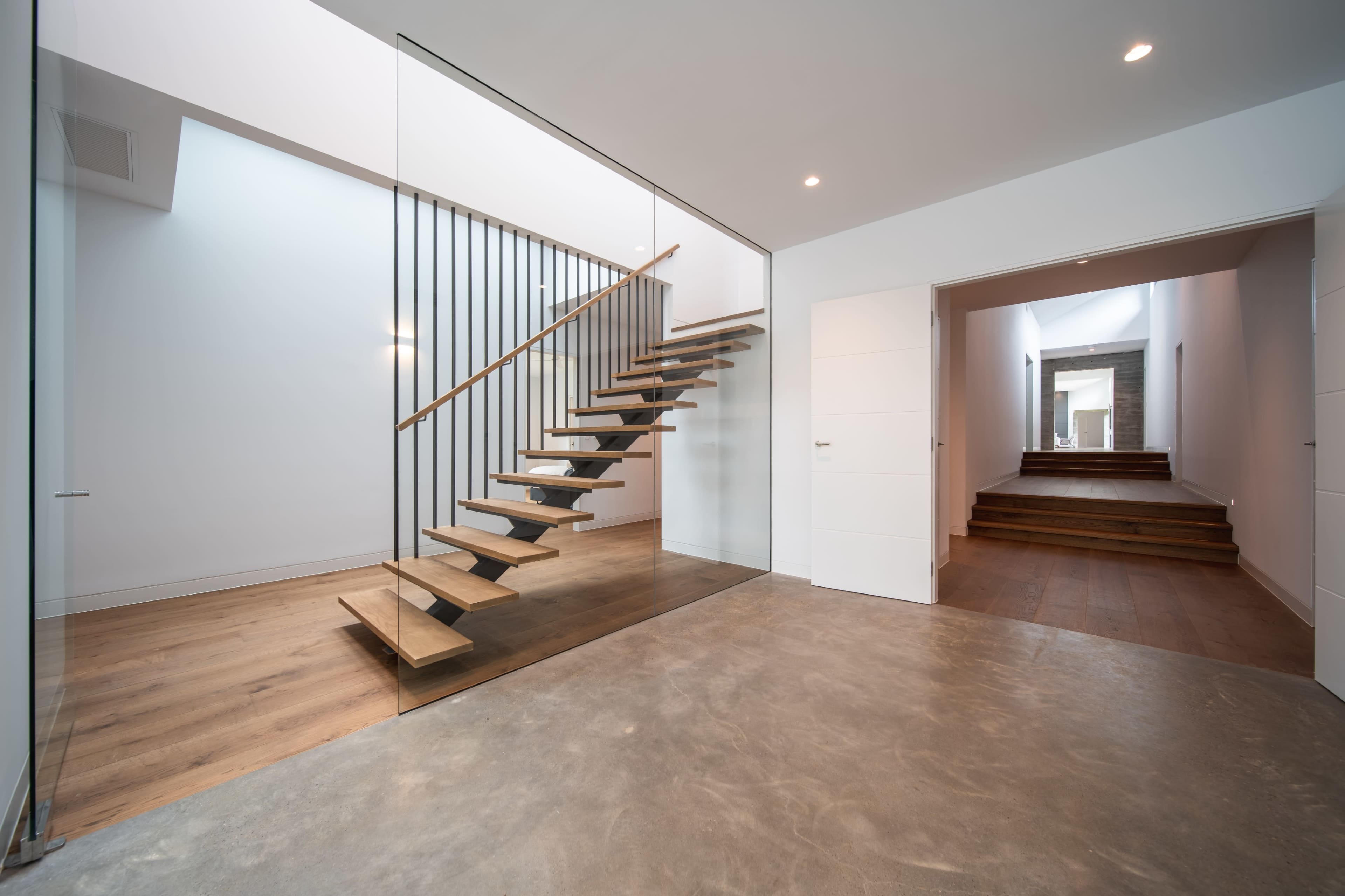 A modern interior space featuring a glass-enclosed staircase made of wooden steps, leading to an upper level, with a hallway visible in the background.