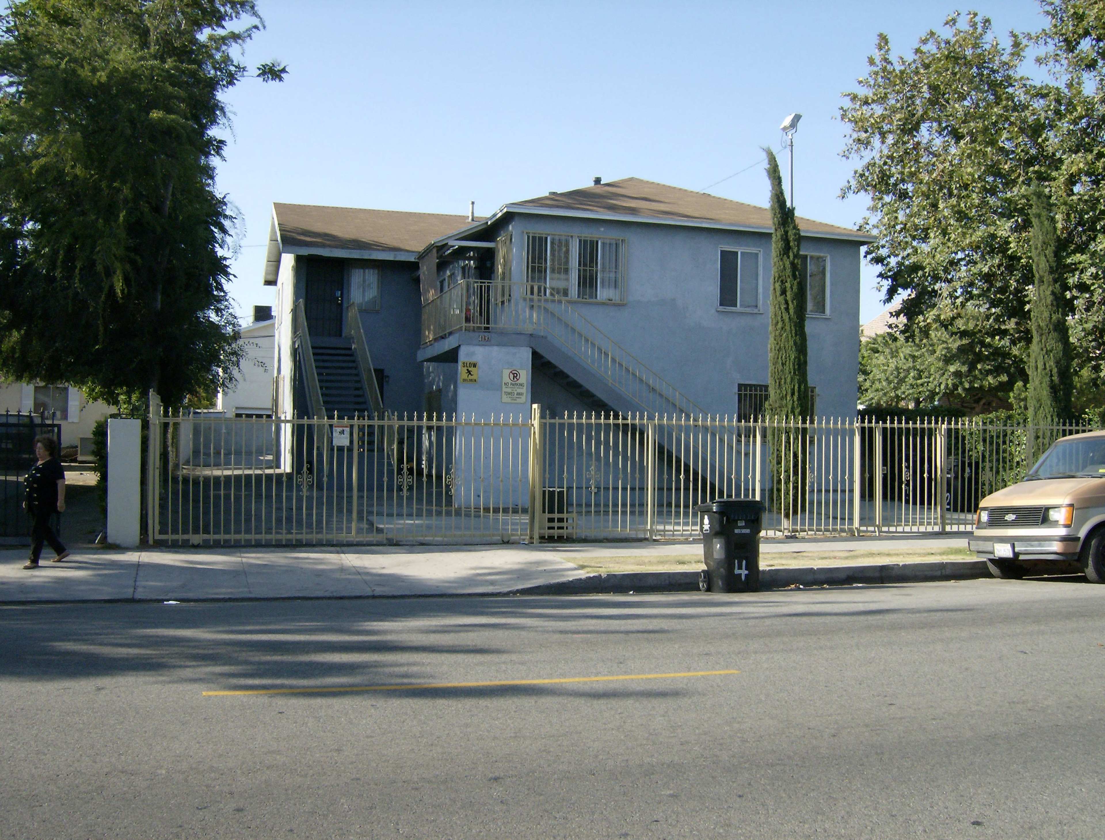 The image shows a two-story blue residential building with a fenced yard and a staircase on the side, located along a street.