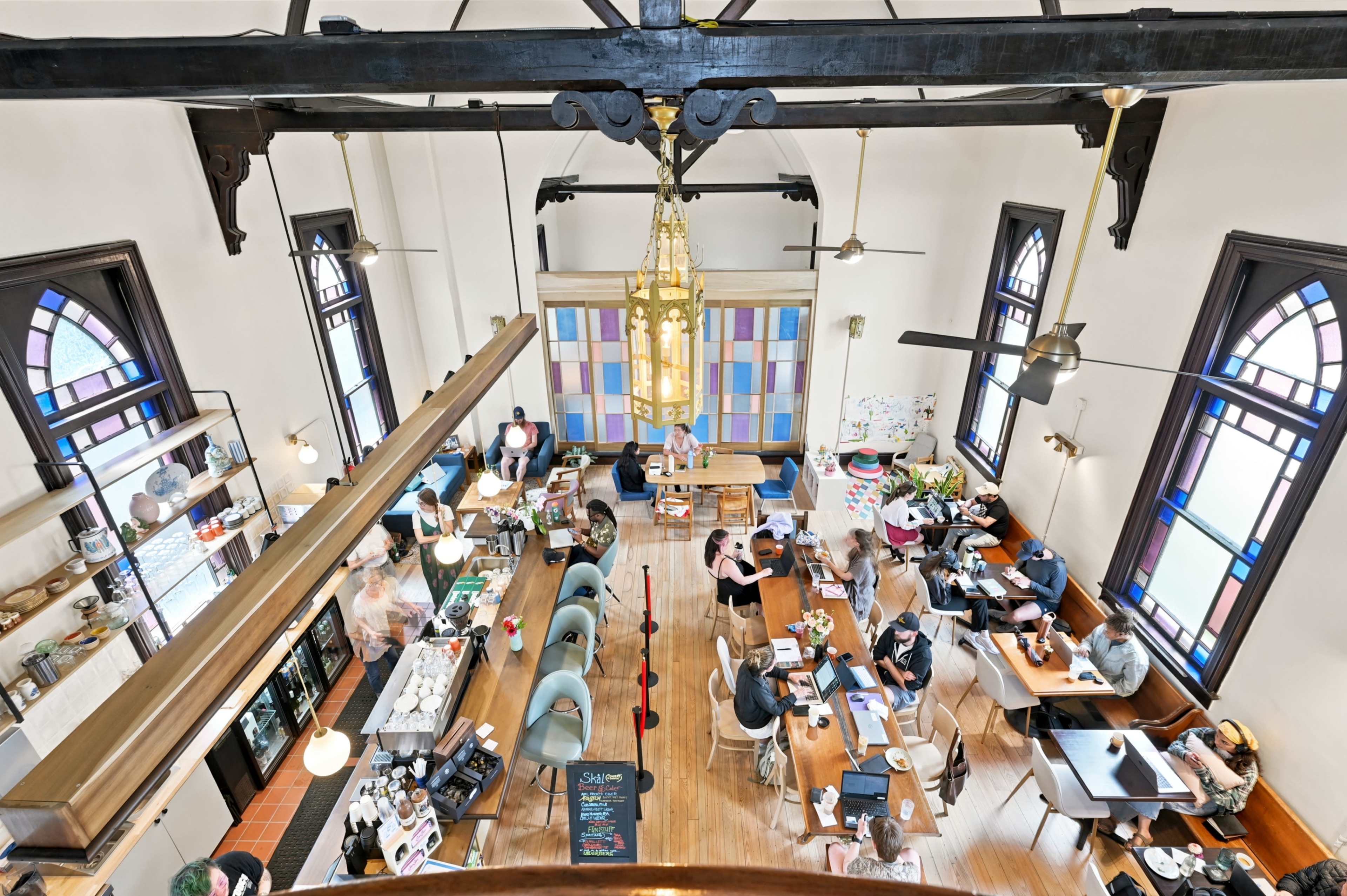 The image shows an overhead view of a bustling café with patrons seated at various tables and a counter displaying food and drinks, all set within an architecturally interesting space featuring tall windows and decorative elements.