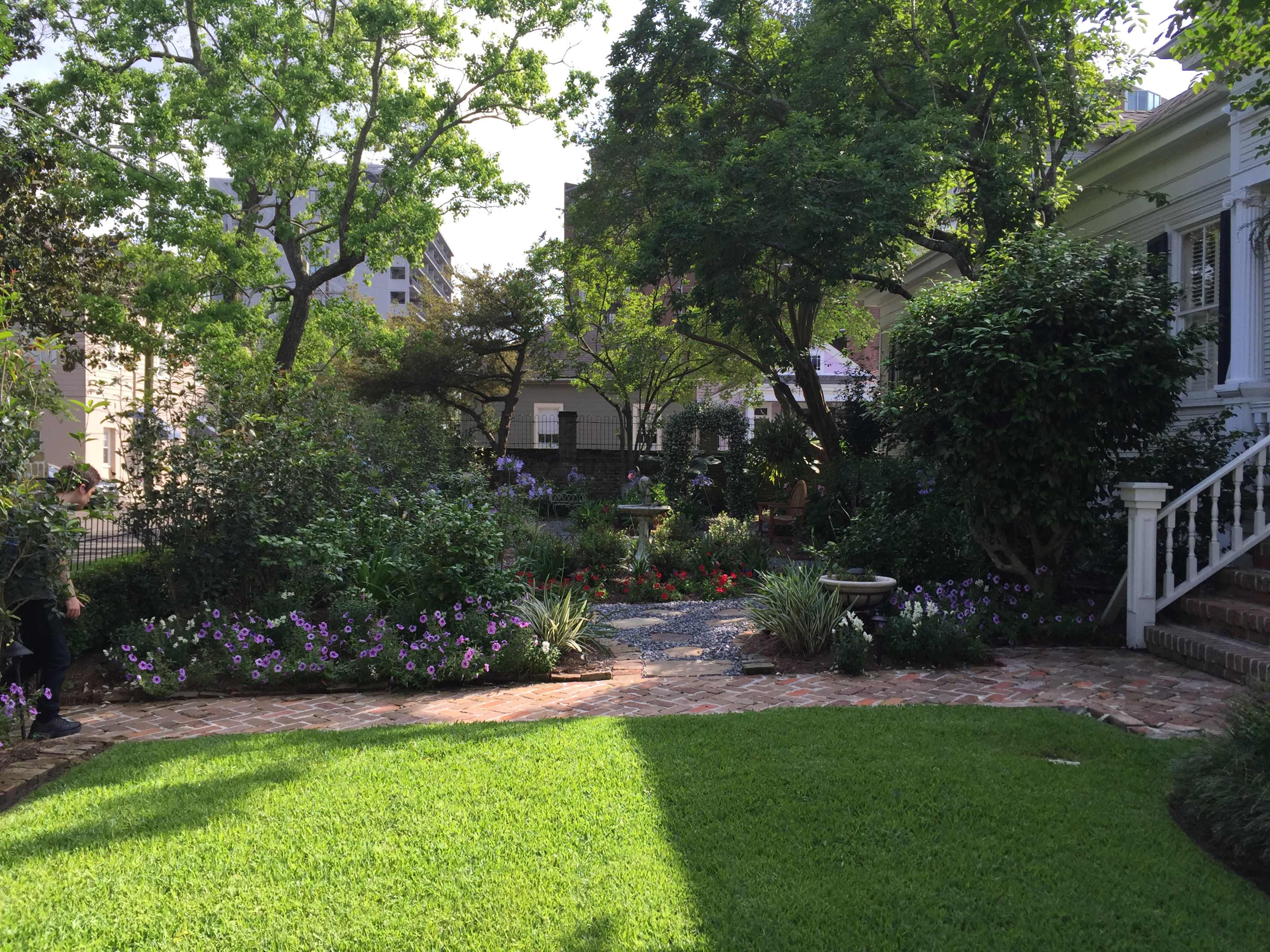 The image shows a landscaped garden with various plants and flowers surrounding a stone pathway, adjacent to a house.