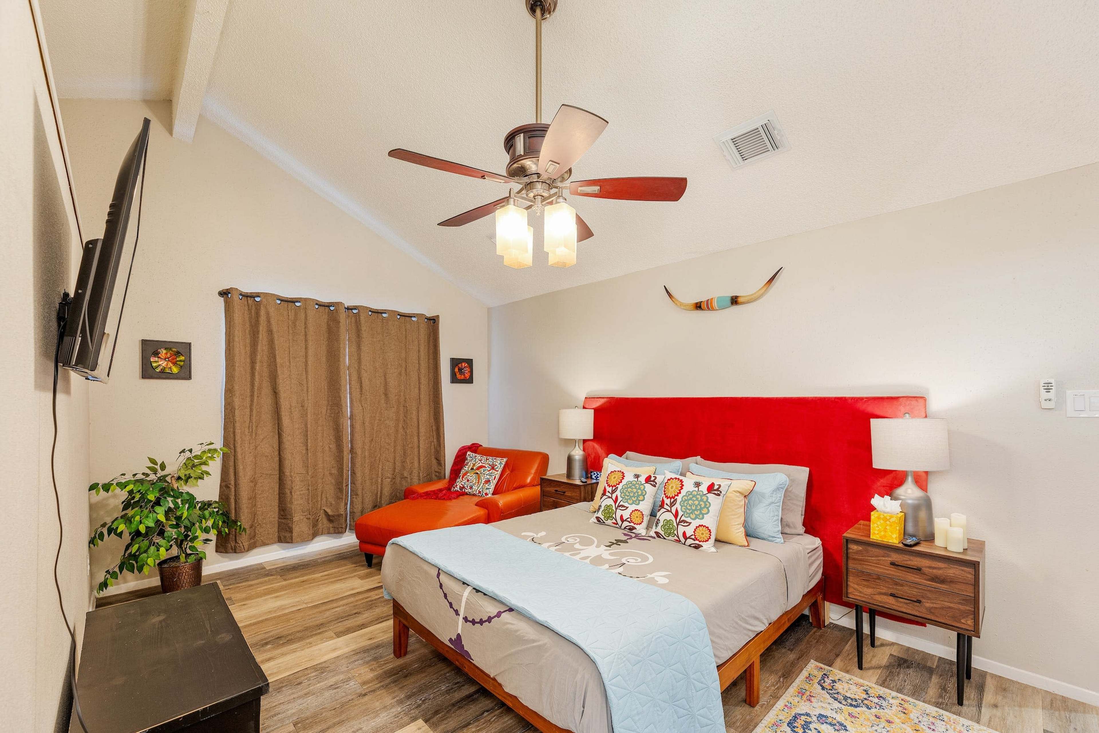 The image shows a well-decorated bedroom featuring a red headboard, a ceiling fan, and a cozy reading corner with an orange chair.