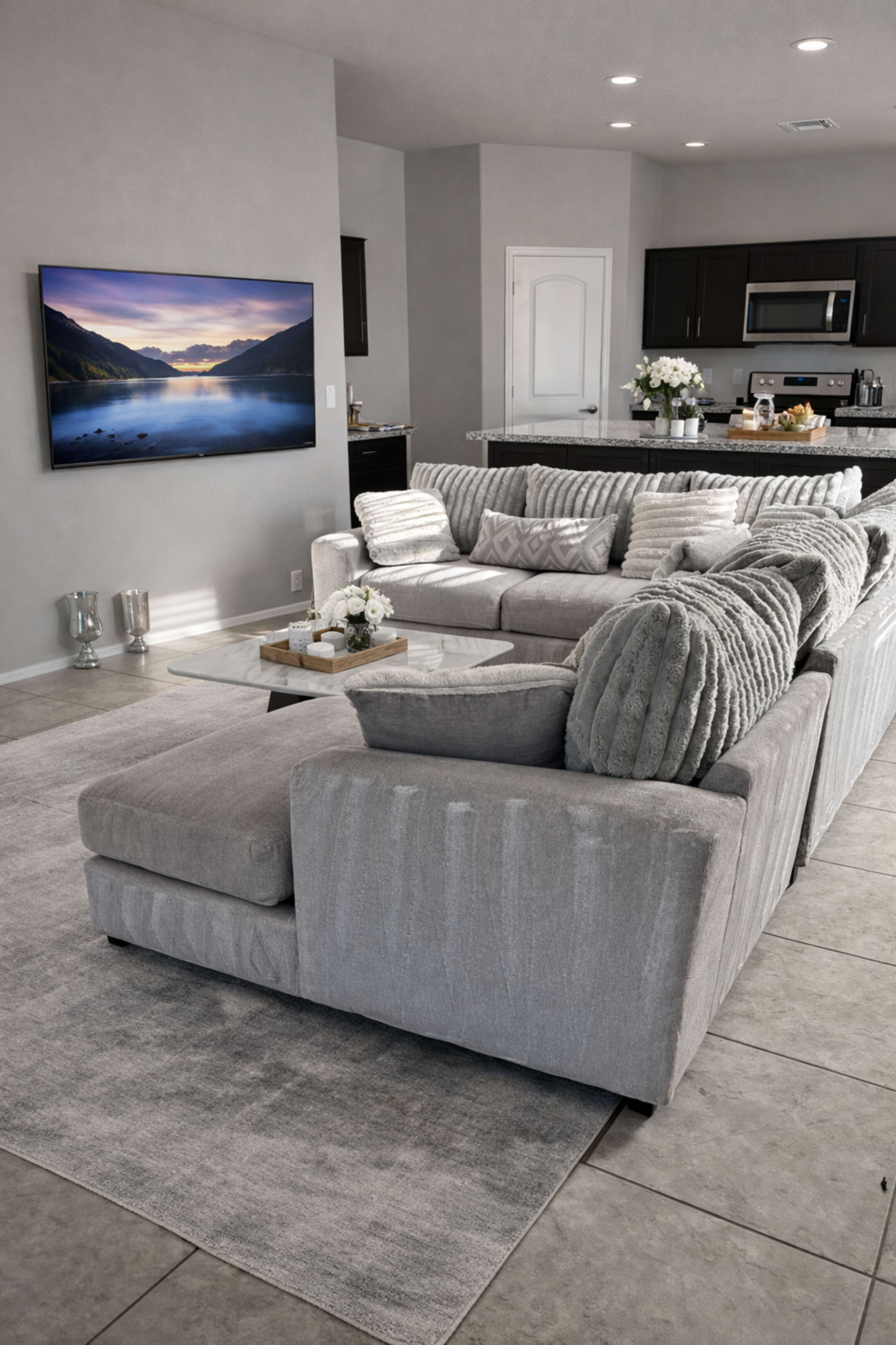 The image shows a modern living room with a large gray sectional sofa, a light gray area rug, and a wall-mounted TV above a kitchen area featuring dark cabinets and a countertop.