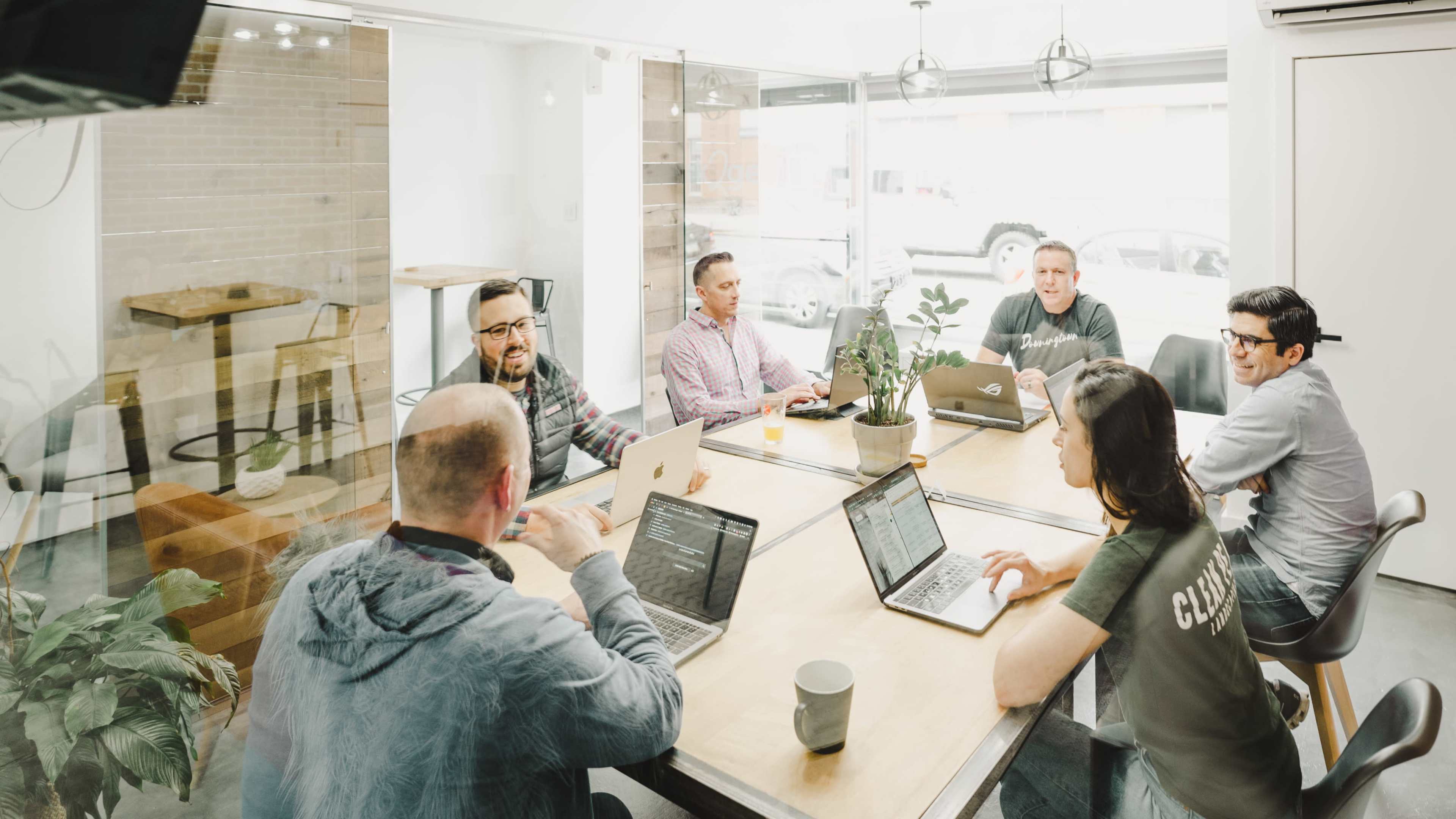A group of six people is seated around a wooden table in a bright meeting room, working on laptops and discussing ideas.