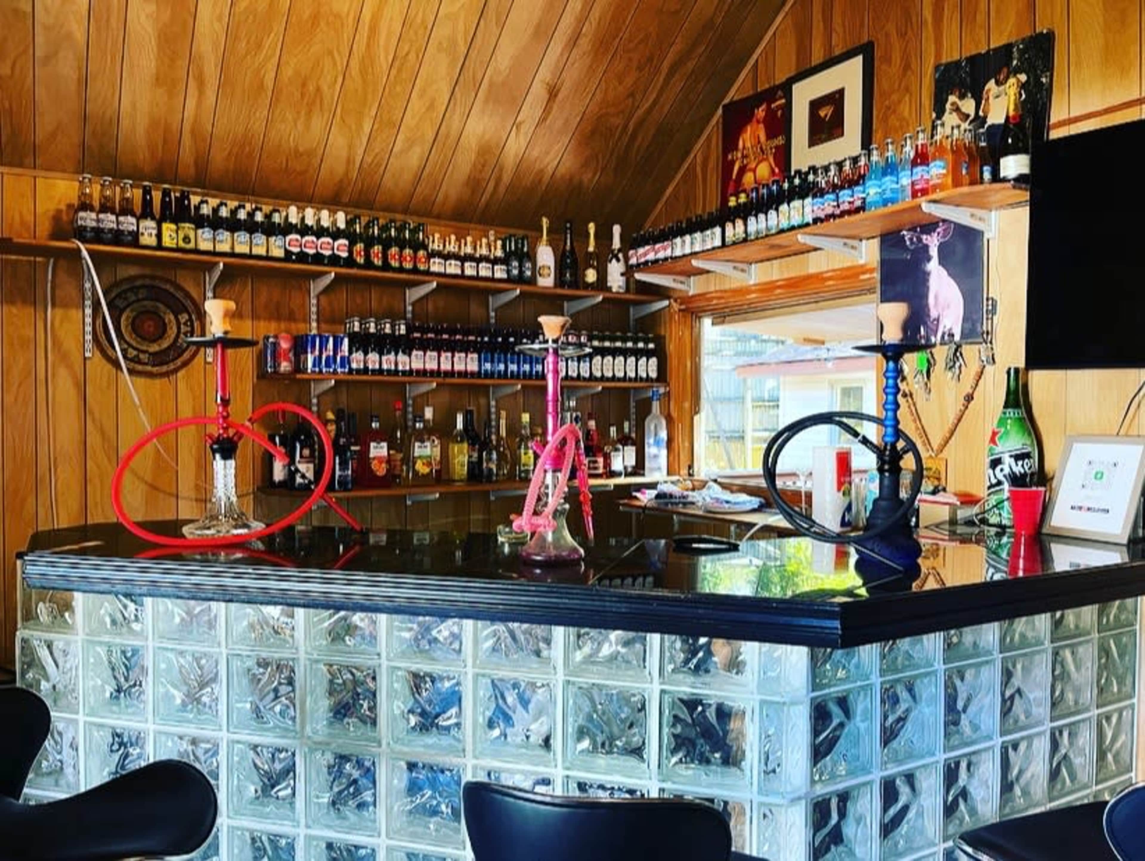 The image shows a home bar with a wooden interior, glass block countertop, various bottles arranged on shelves, and two hookahs on the bar surface.