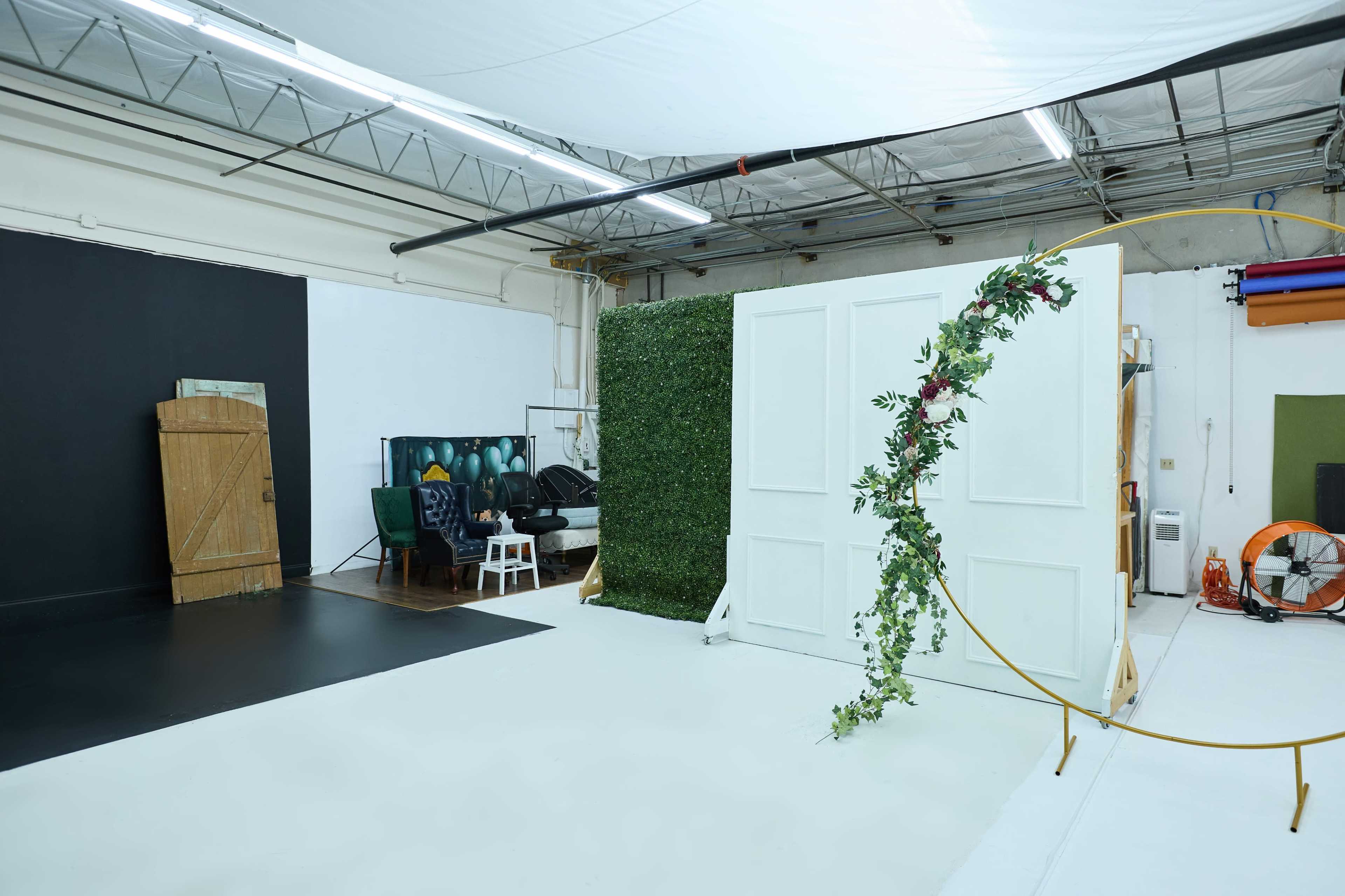 A spacious studio with a black backdrop, a white partition adorned with greenery and flowers, and various furniture pieces arranged around the area.