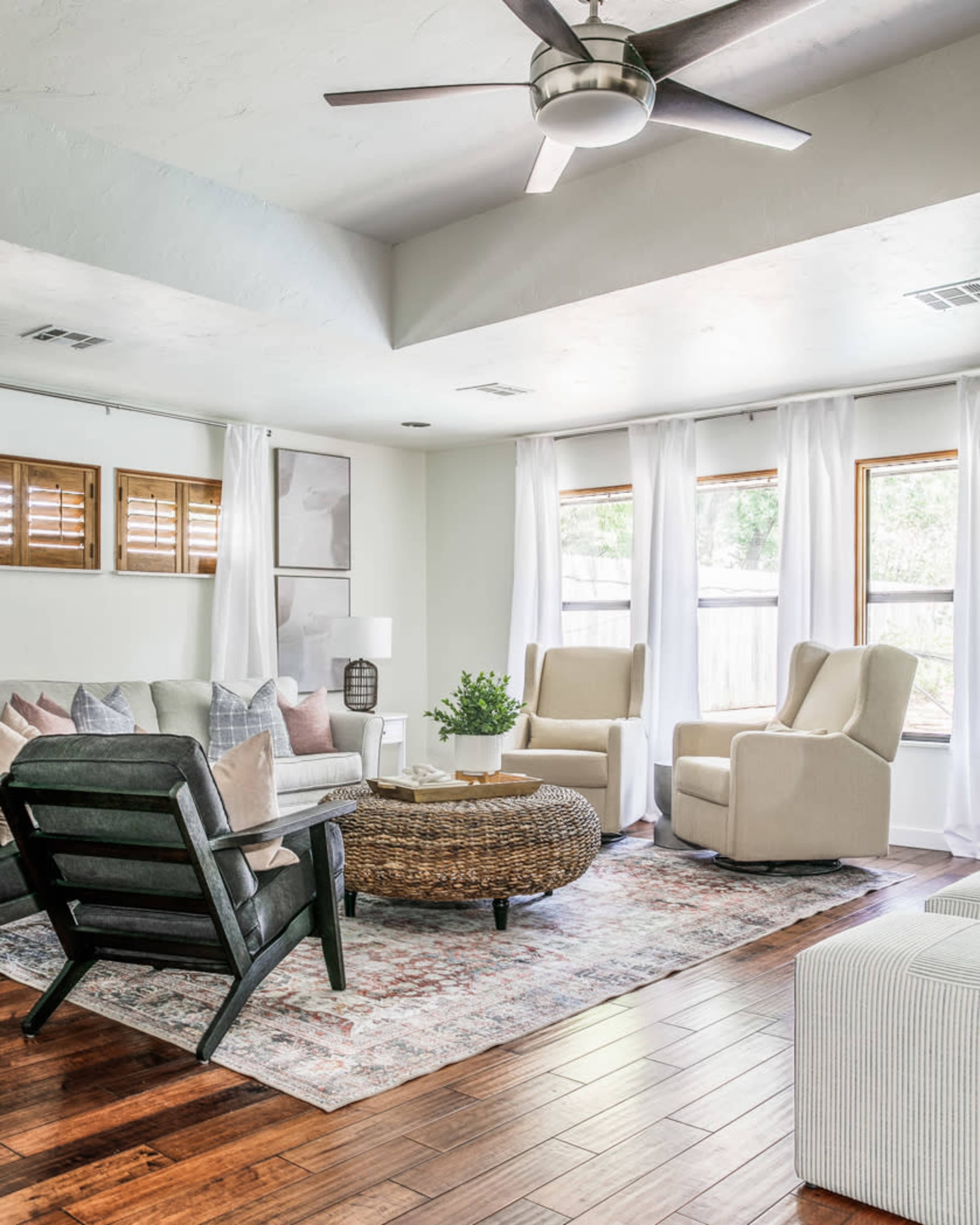 A living room features two armchairs, a round coffee table, a sofa, and large windows with sheer curtains, all set on a hardwood floor.