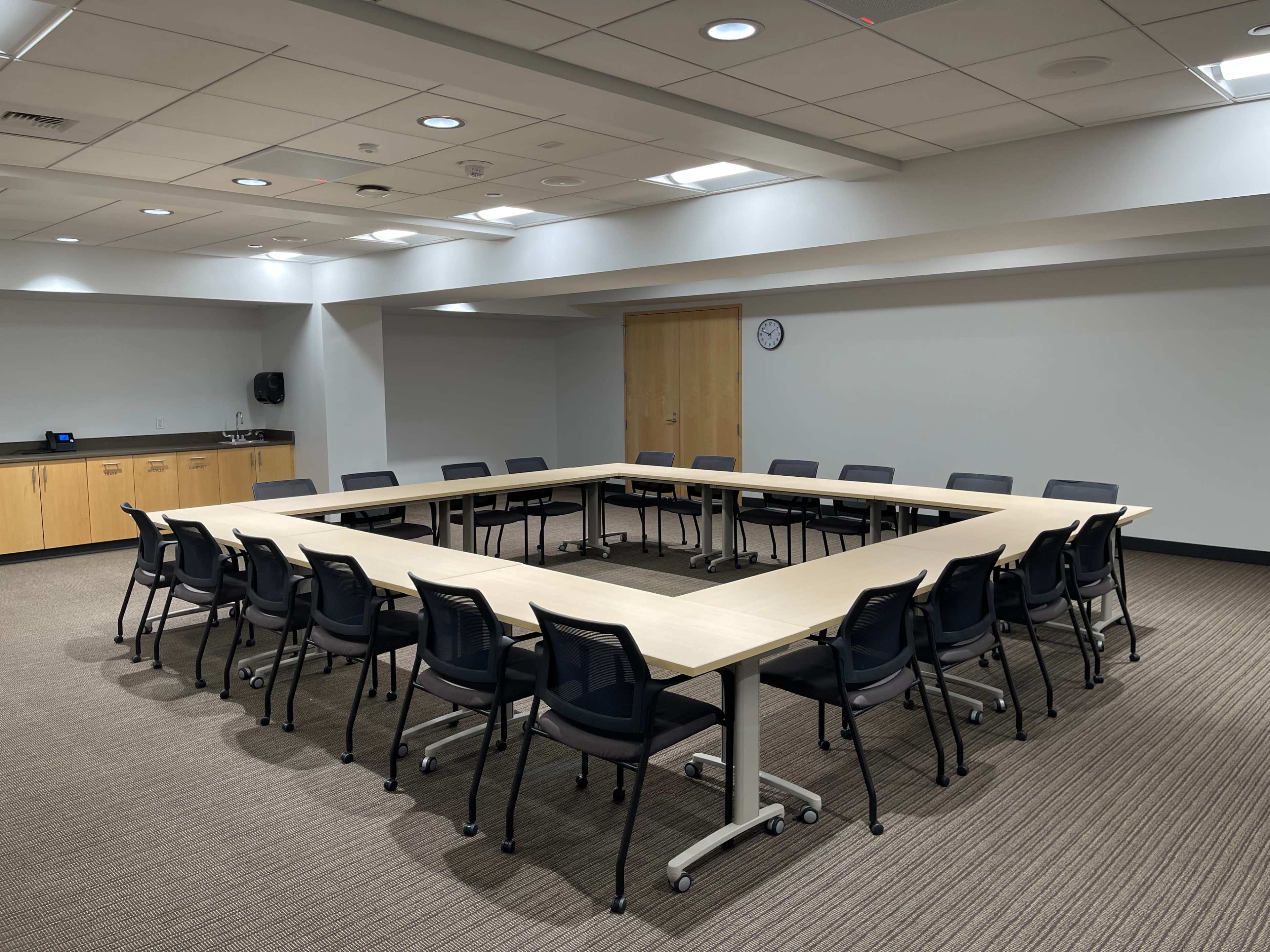 A large conference room features a long table arranged in an open square shape, surrounded by black chairs, with wooden cabinets and a clock visible on the walls.