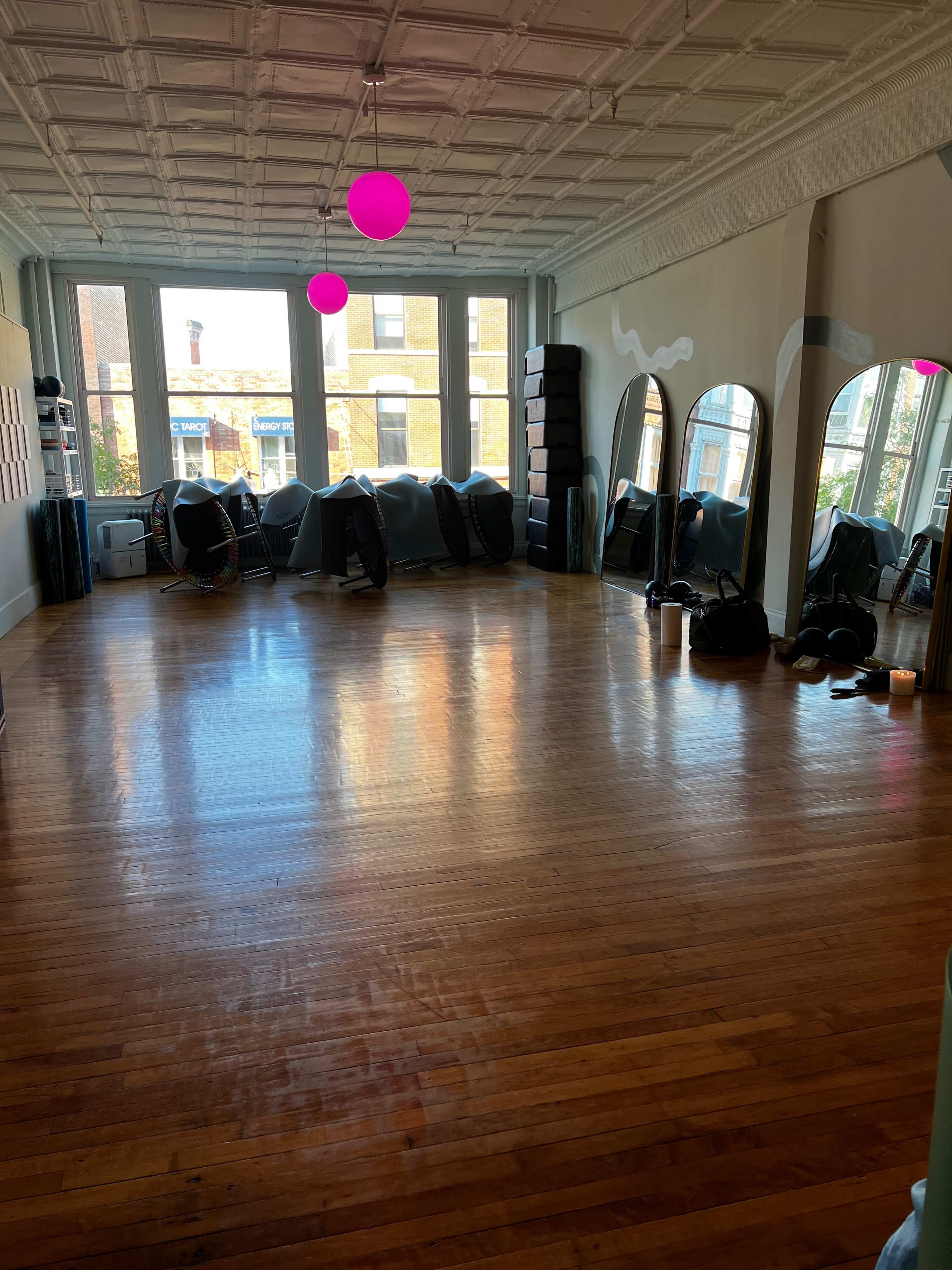 The image shows a spacious, empty studio with wooden floors, large windows, and mirrored walls, illuminated by pink pendant lights.