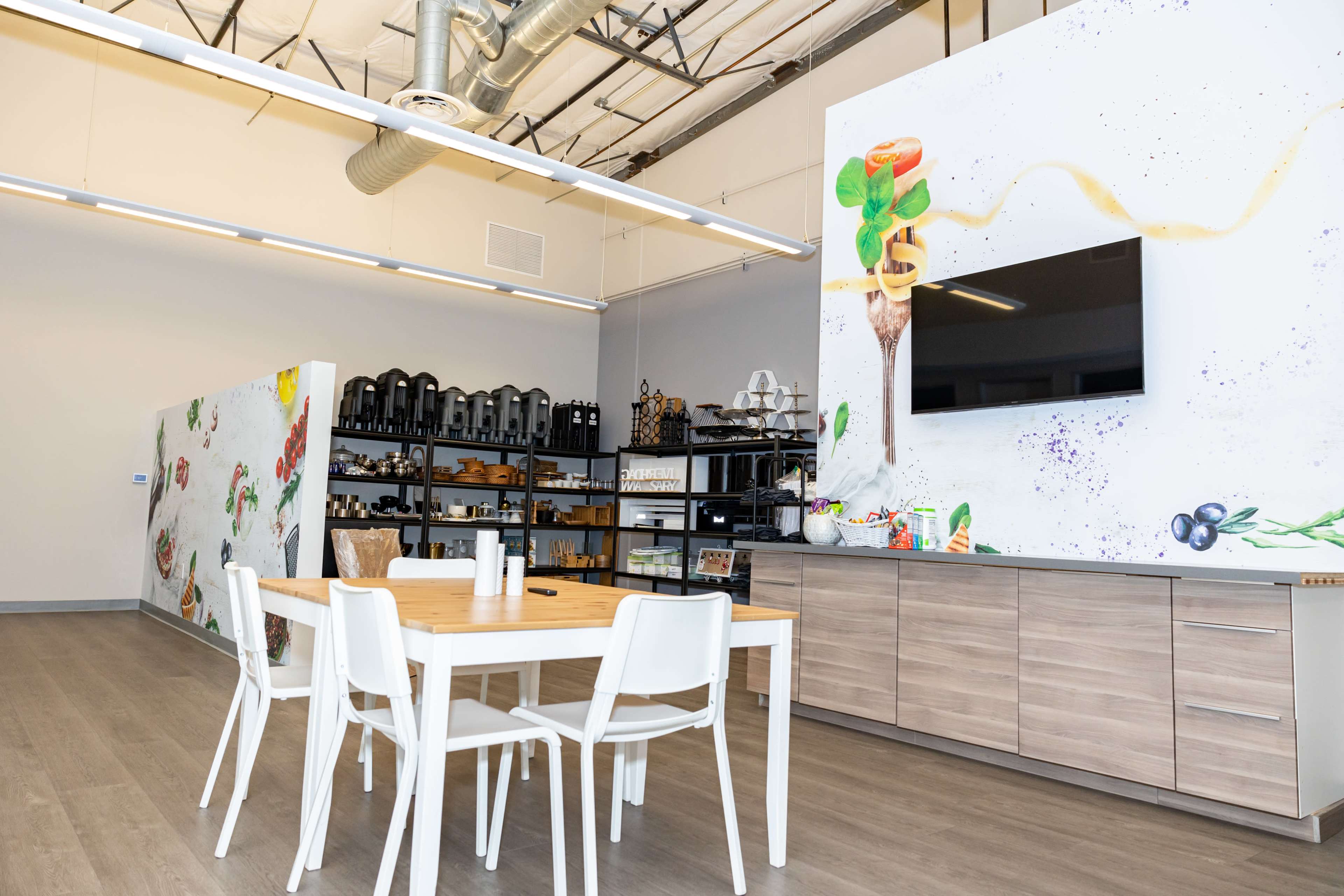 The image shows a modern kitchen space featuring a wooden dining table with white chairs and colorful wall murals alongside shelves filled with kitchenware.