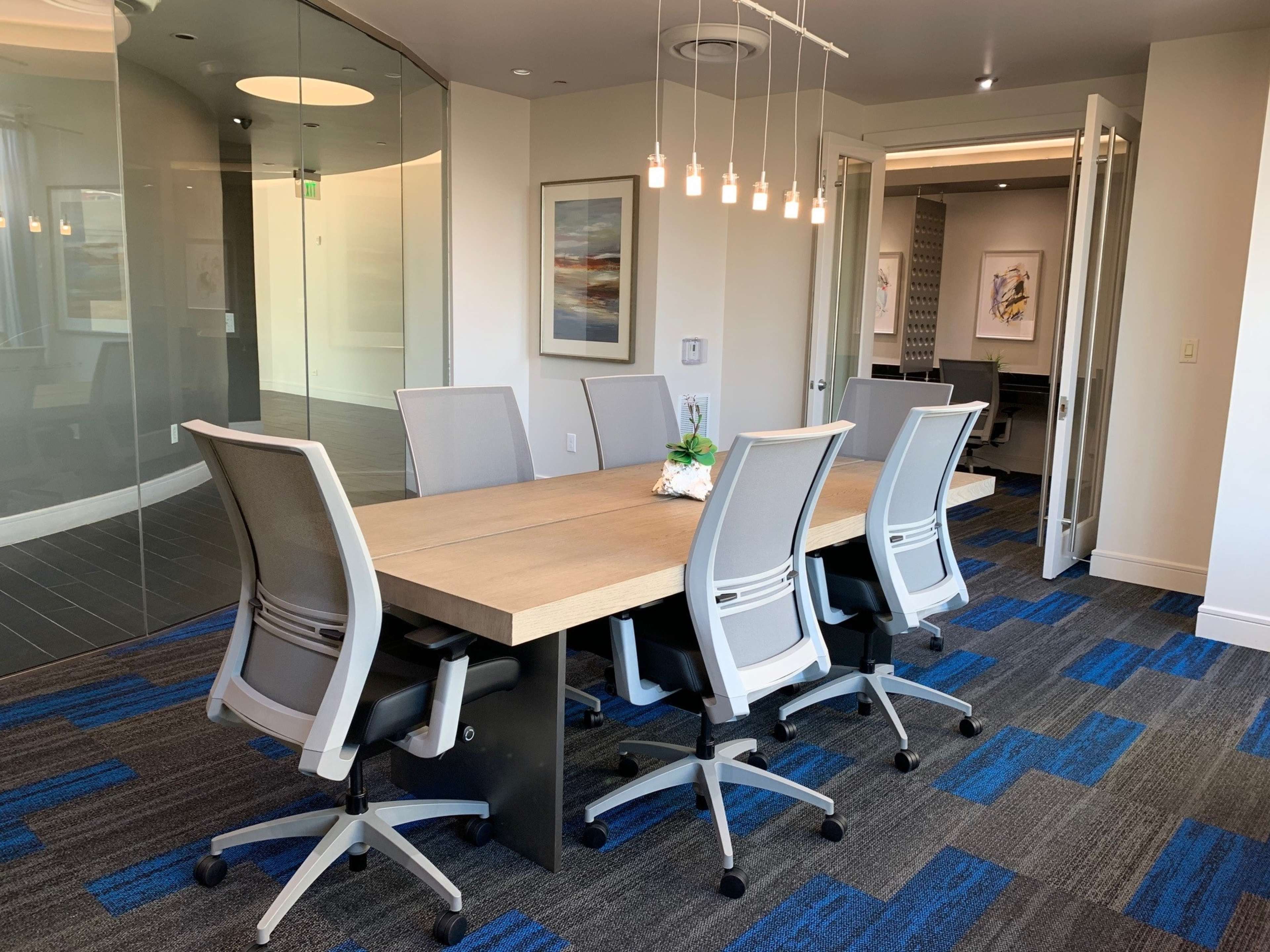 A modern conference room features a long table surrounded by six ergonomic chairs, with a glass wall and hanging light fixtures above.