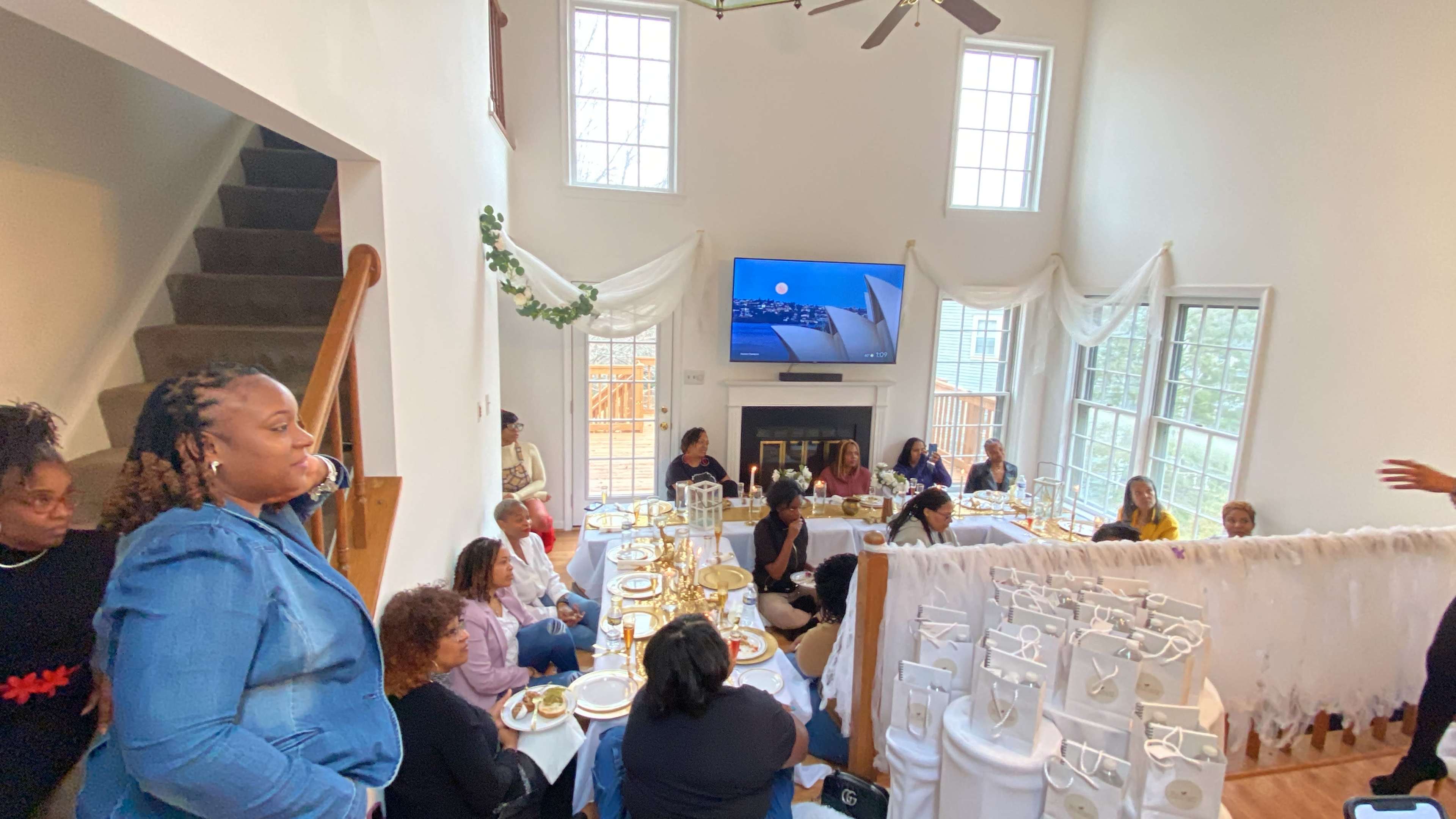 A group of people gathers in a well-lit living room, seated around tables set for a meal, while a large television displays a scenic image.