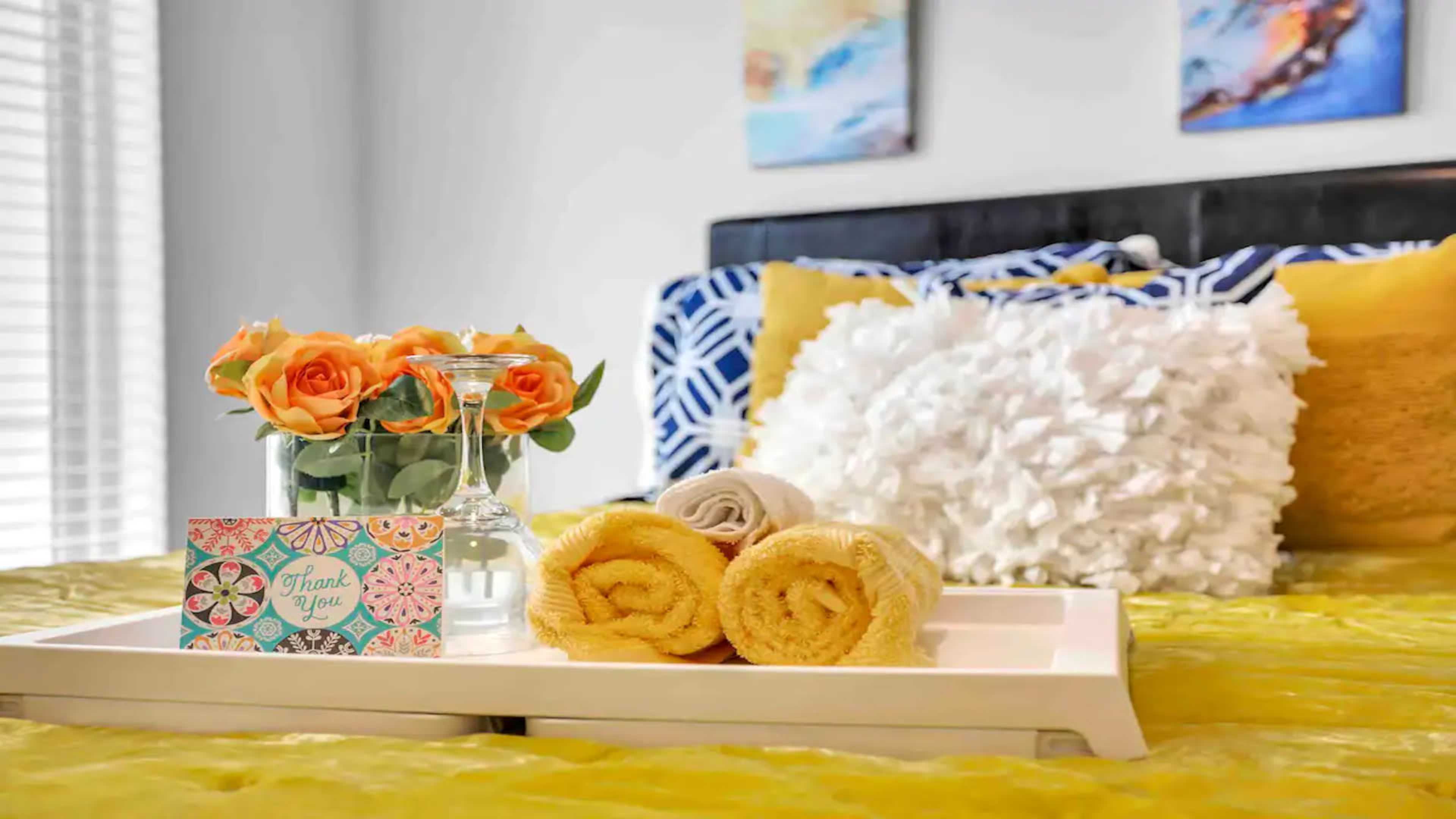 A neatly arranged tray holds a vase of orange roses, a small bottle of water, and rolled towels on a yellow bedding set in a decorated bedroom.