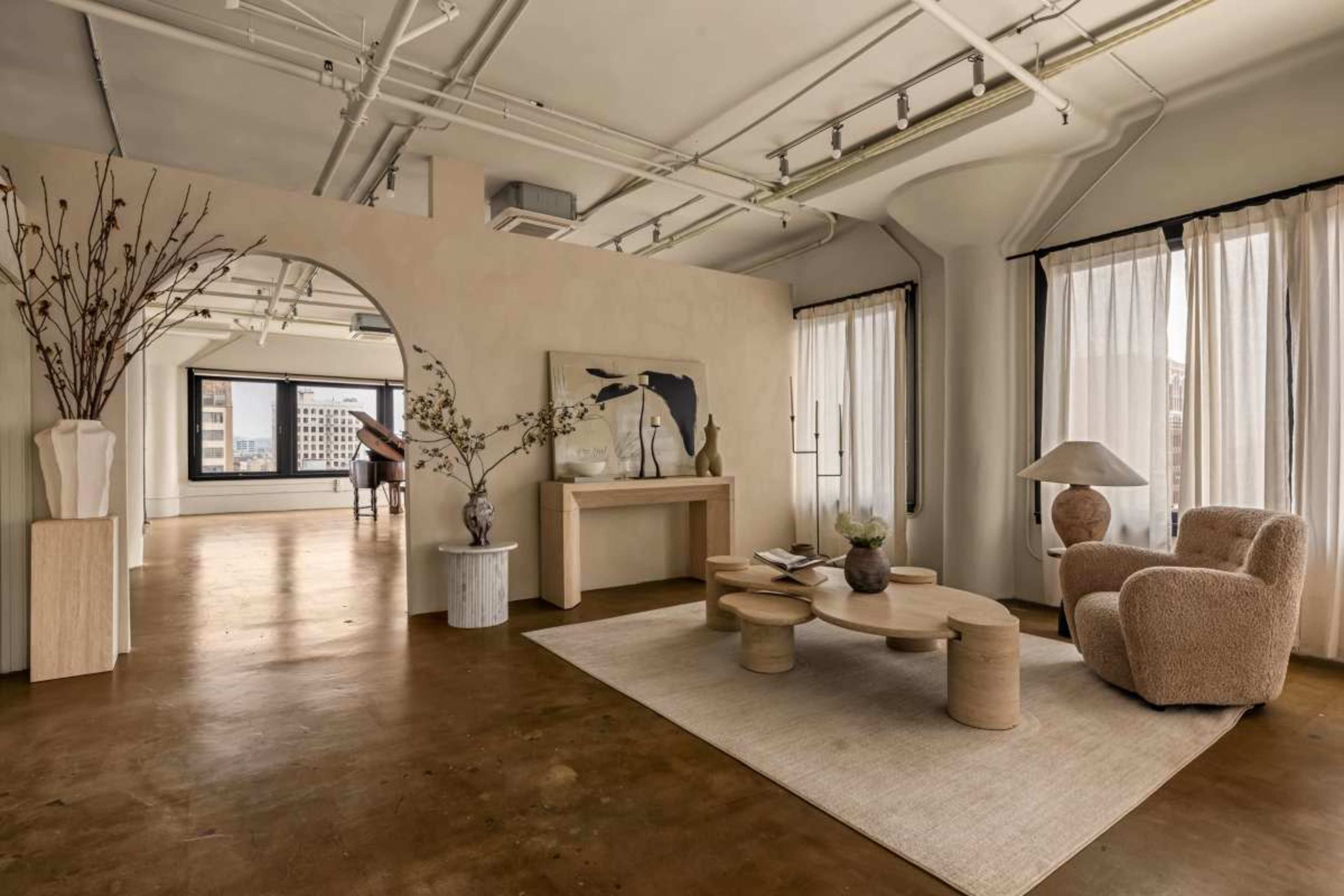 The image shows a modern, minimalist living space with natural light, featuring a round coffee table, a plush armchair, decorative branches, and large windows.