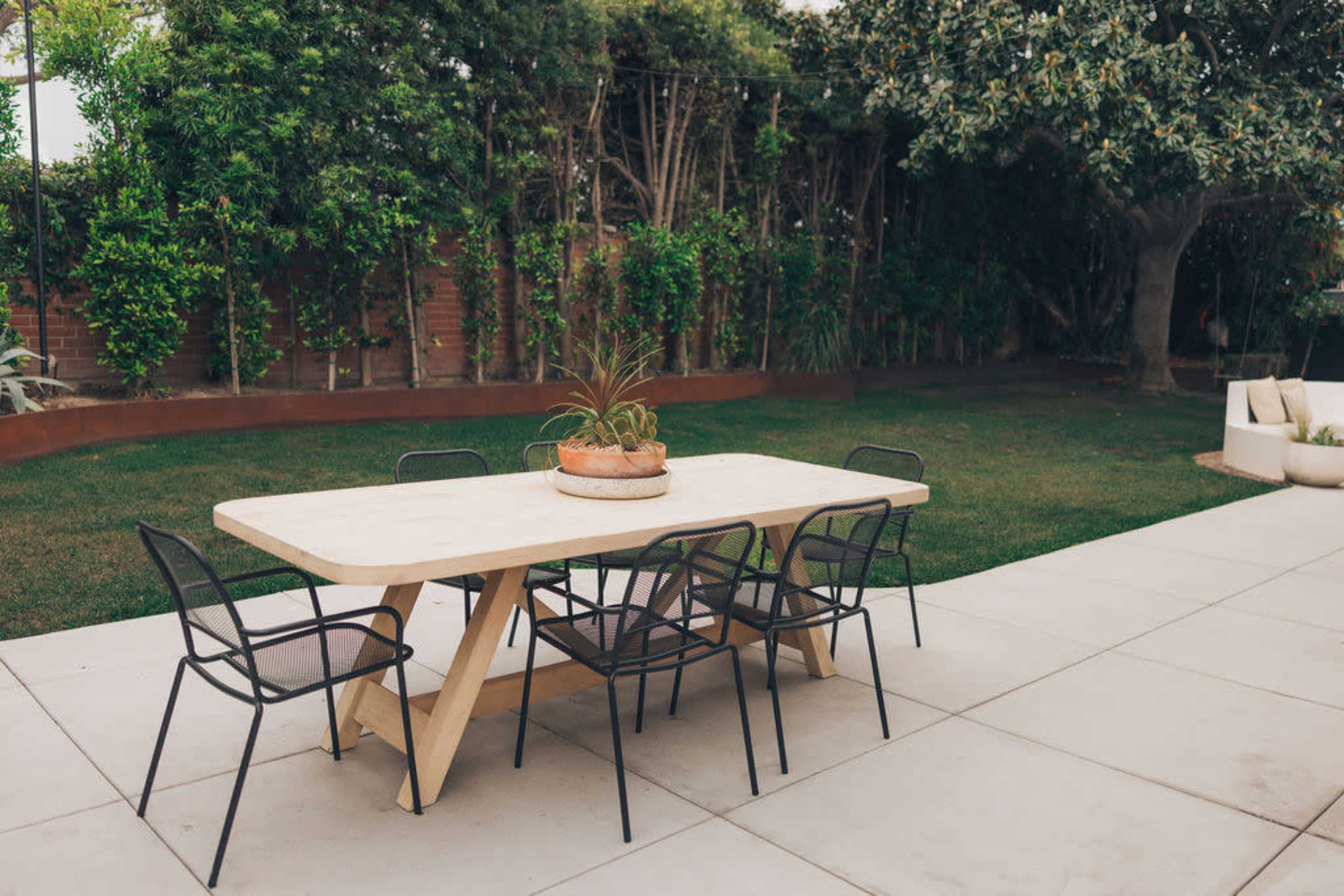A dining table with six black chairs is set on a concrete patio, surrounded by a green lawn and plants in a backyard.