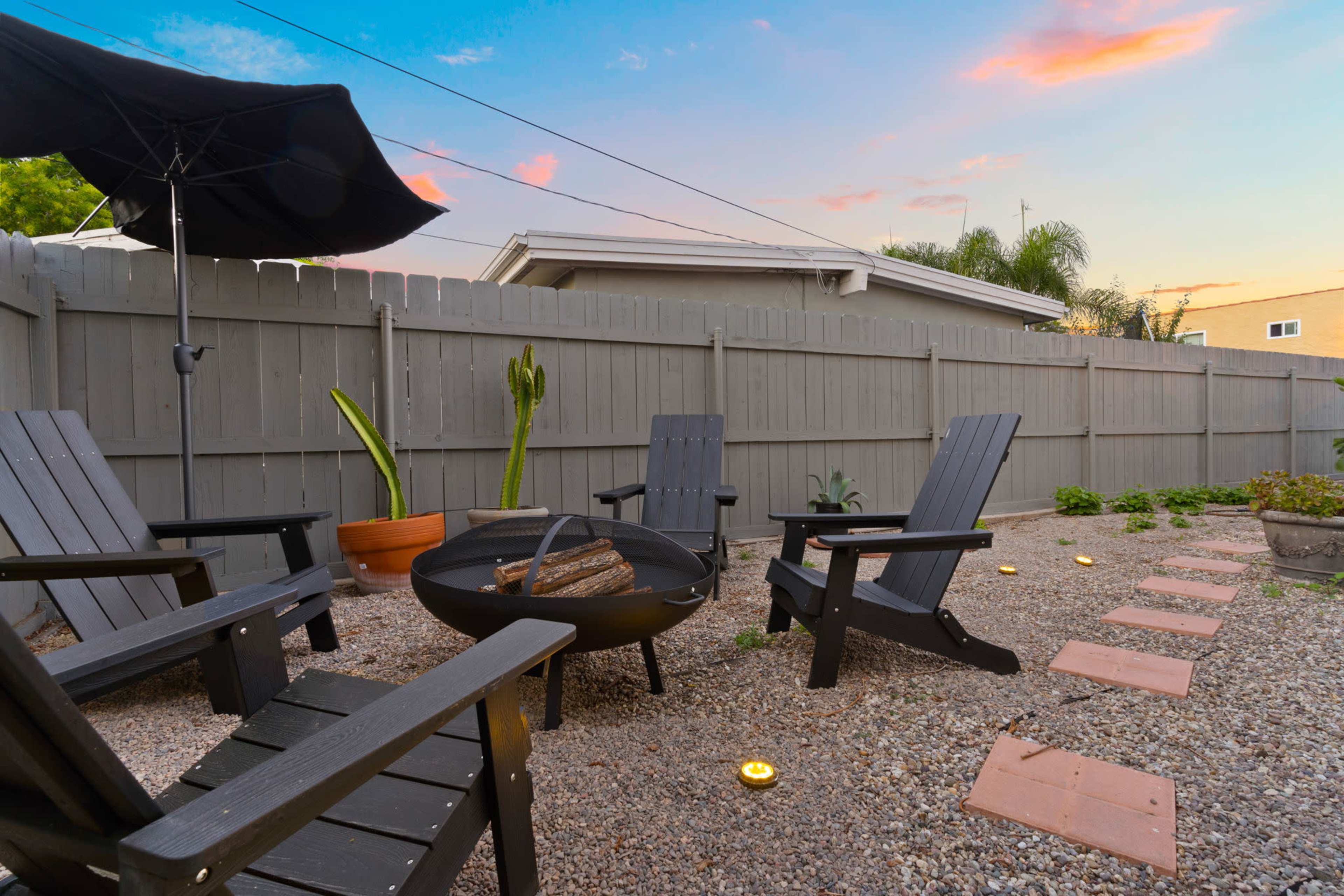 A backyard patio with four black adirondack chairs surrounding a fire pit, stone pathways, and potted plants against a wooden fence.