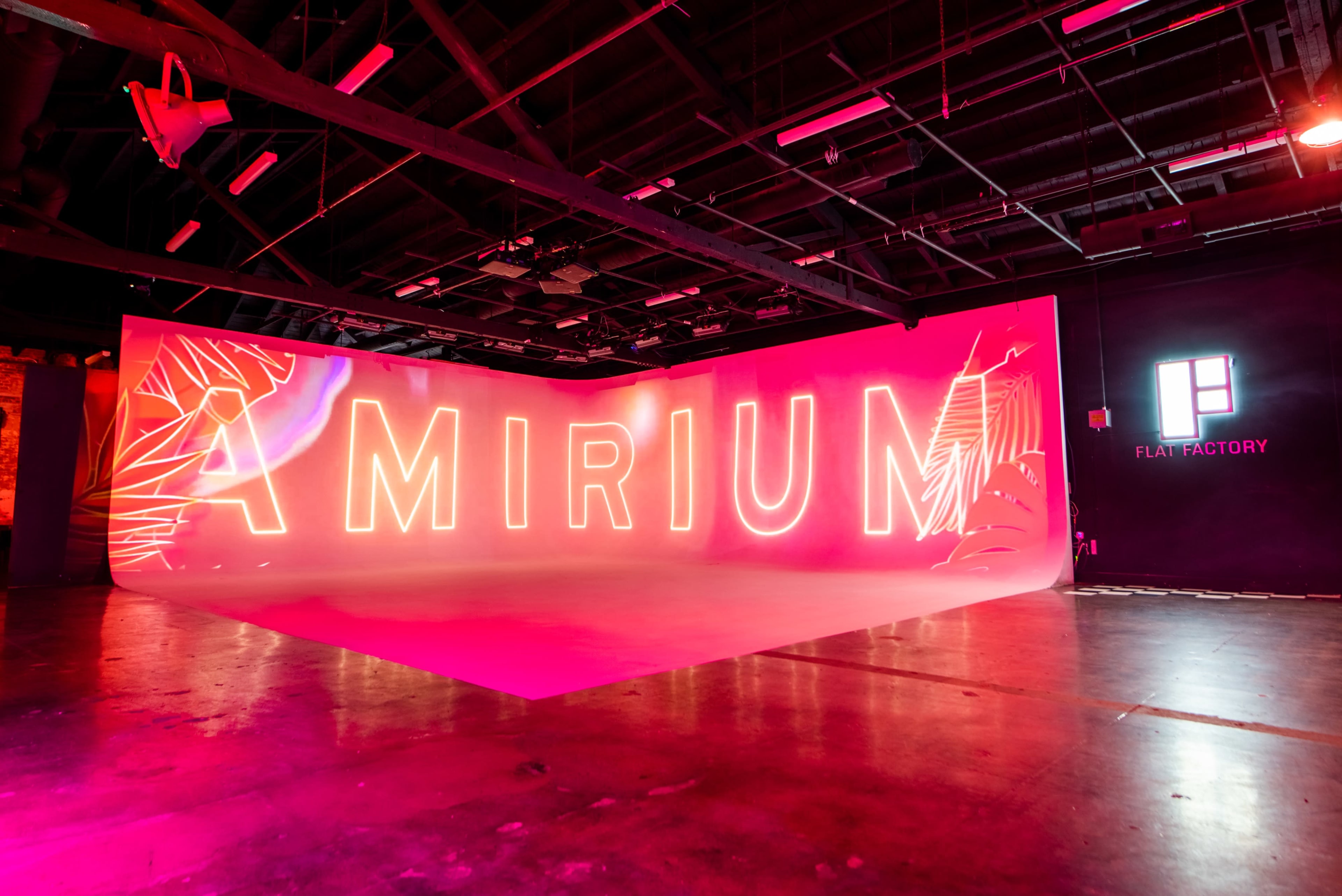 The image shows a brightly lit backdrop featuring the word "AMIRIUM" in large, glowing letters, set in an industrial-style space with a pink and white color scheme.