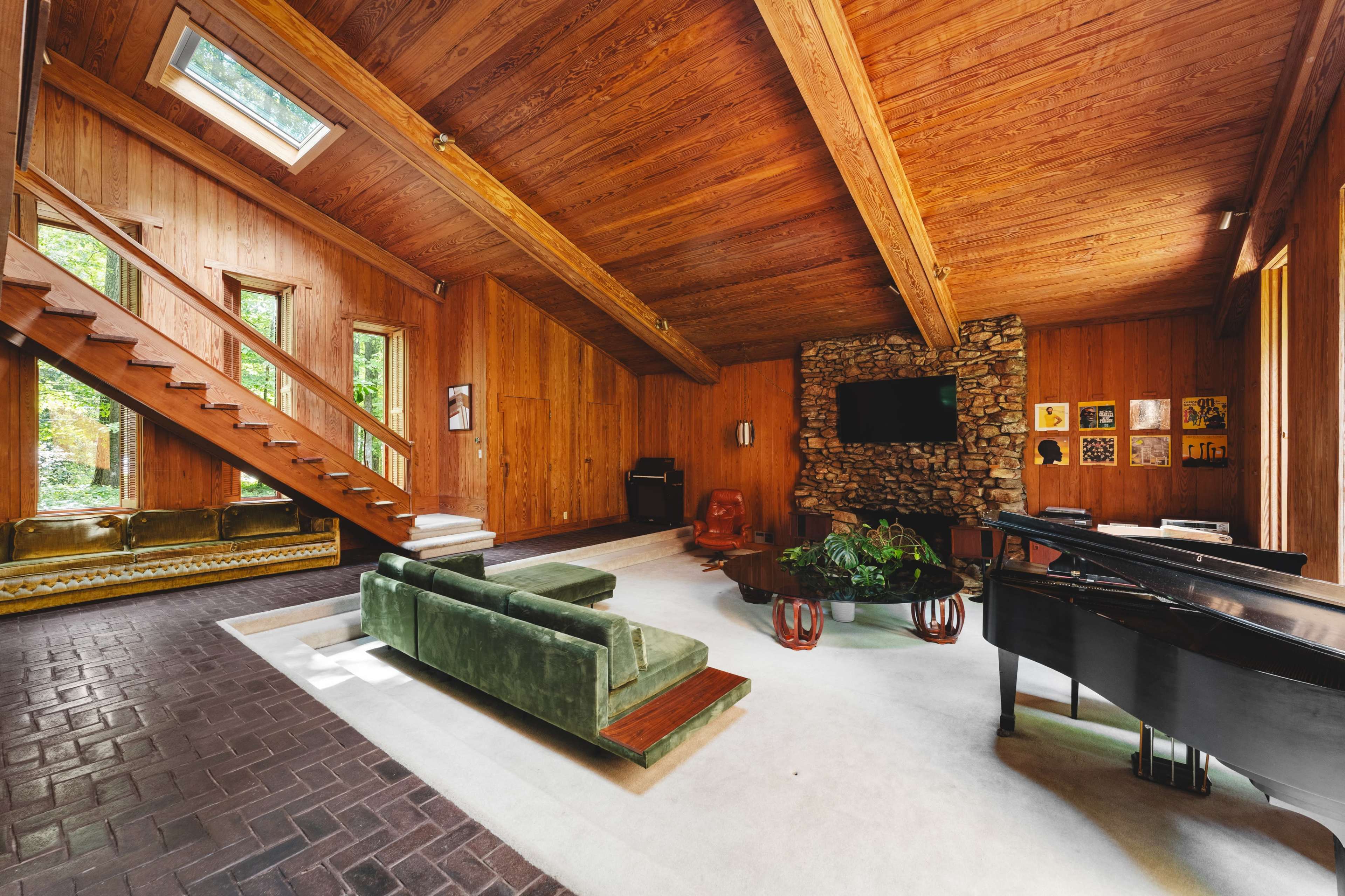 The image depicts a spacious living area featuring wooden walls and ceiling, a green sofa, a black piano, and a stone fireplace, with large windows allowing natural light to enter.