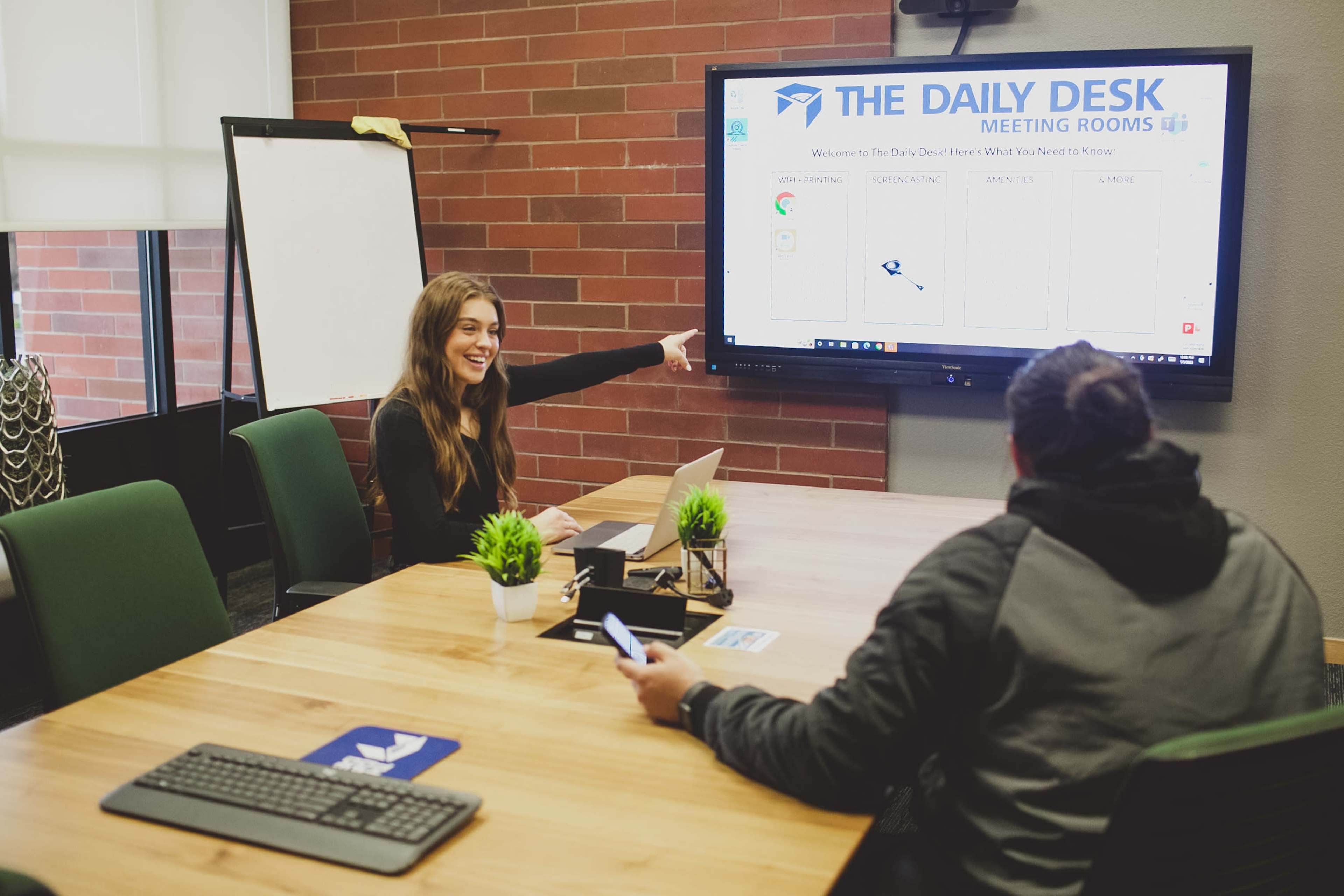 A woman points at a screen displaying information in a meeting room while a man sits across the table, looking at a laptop.