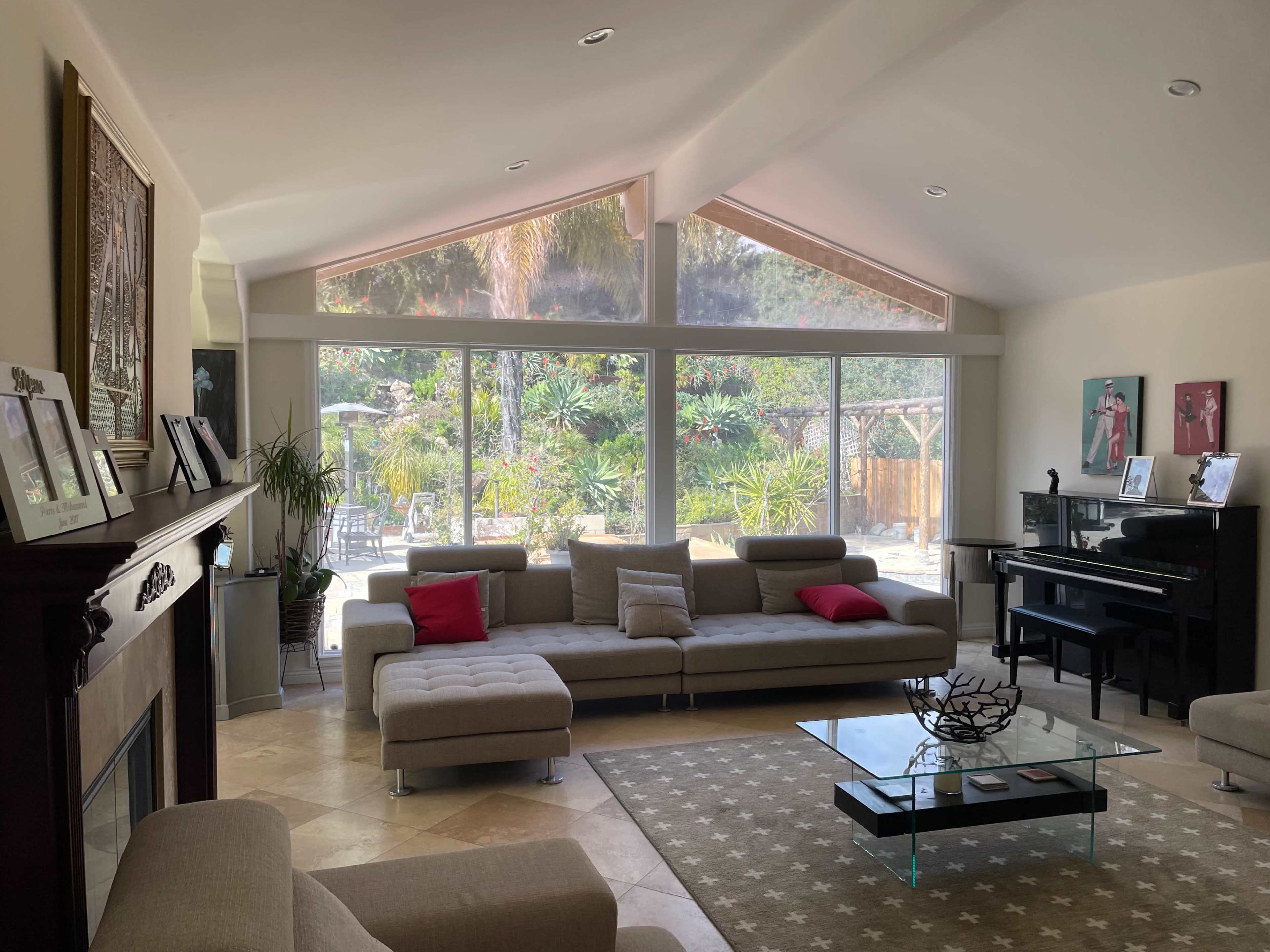 The living room features a large sectional sofa, a coffee table, and a grand piano, with expansive windows overlooking a garden area.