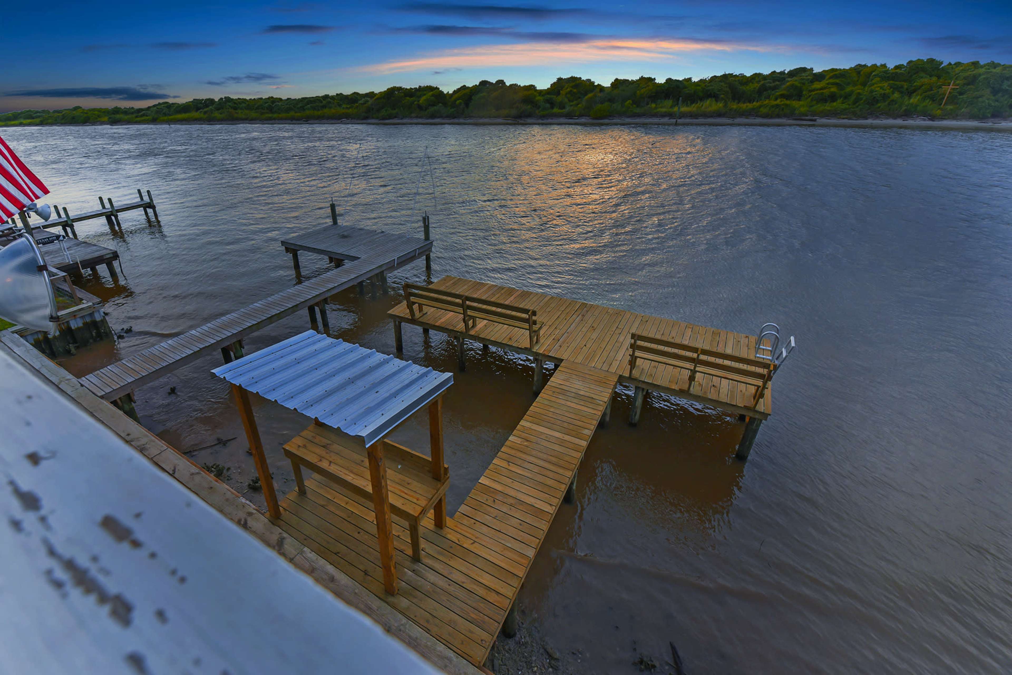 A wooden dock extends into a calm river at sunset, with benches and a shaded area visible.