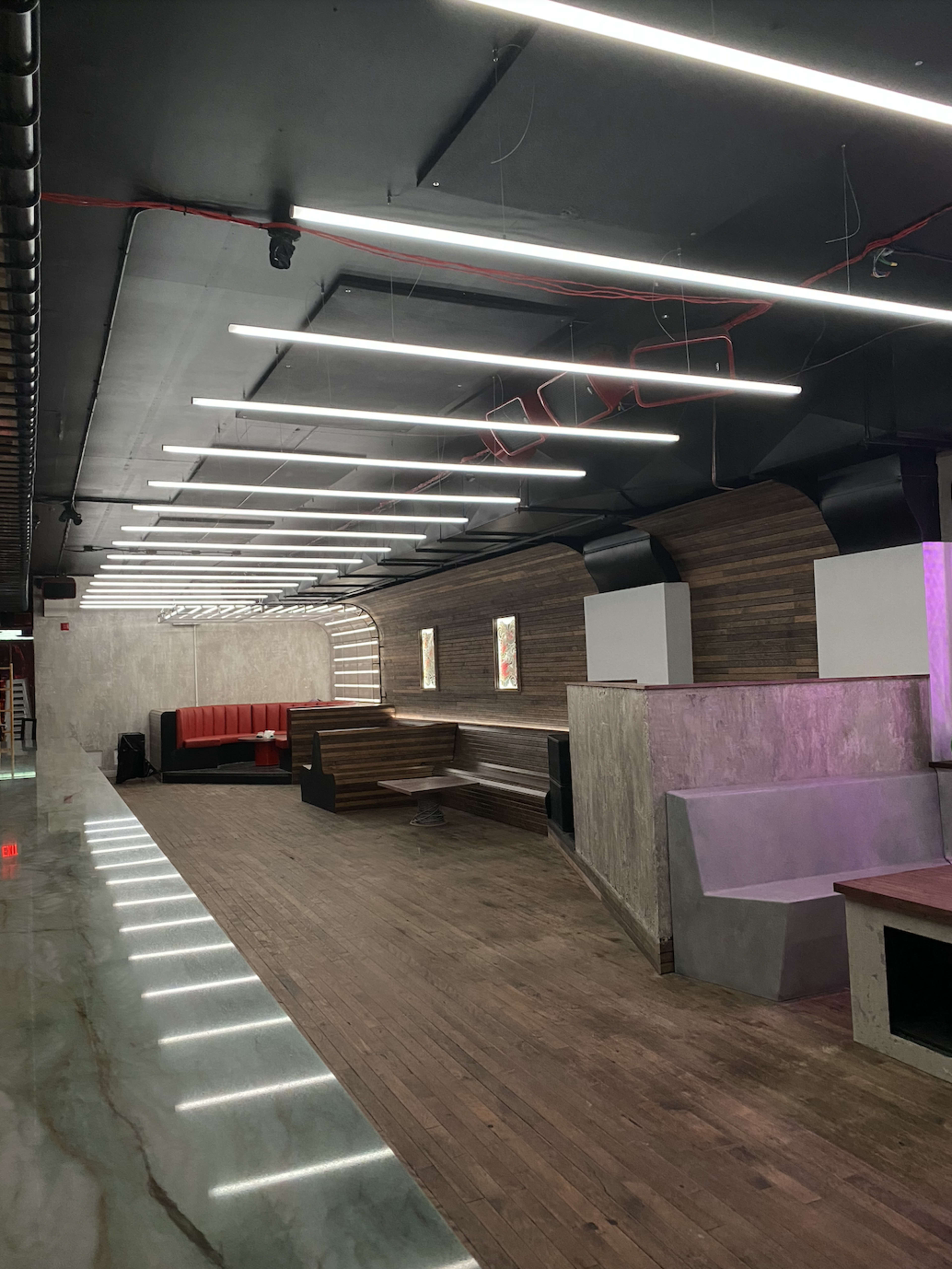 The image shows a modern interior space with wooden seating areas, concrete walls, and overhead lighting.